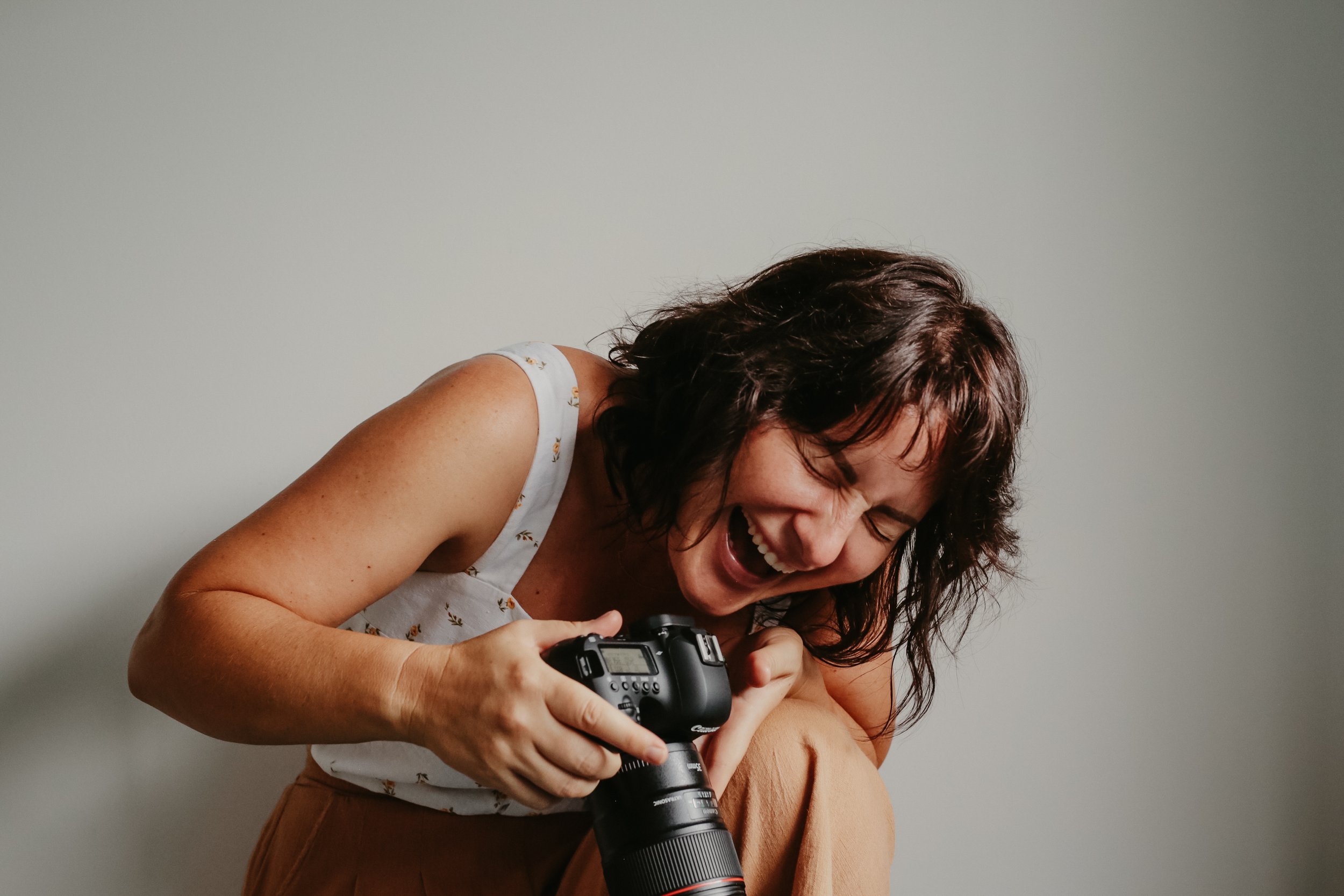A woman with dark brown hair, wearing a white sleeveless top with a small pattern, is laughing joyfully while holding a professional camera and lens. She appears to be in a happy, candid moment. She is a Toowoomba and Brisbane family photographer