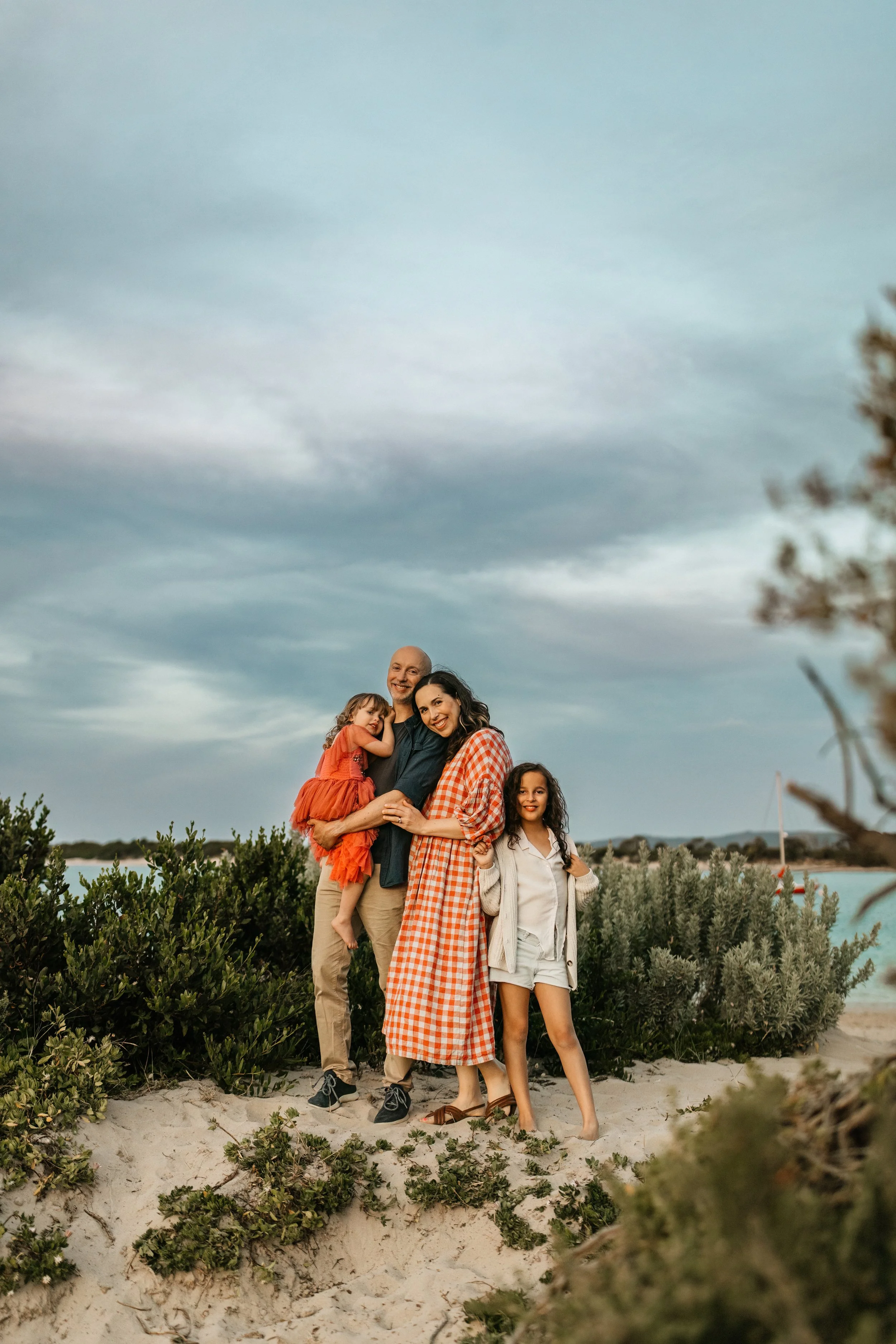 A family of four standing on a sandy beach with green shrubs and bushes, under a cloudy sky, smiling and enjoying each other's company.