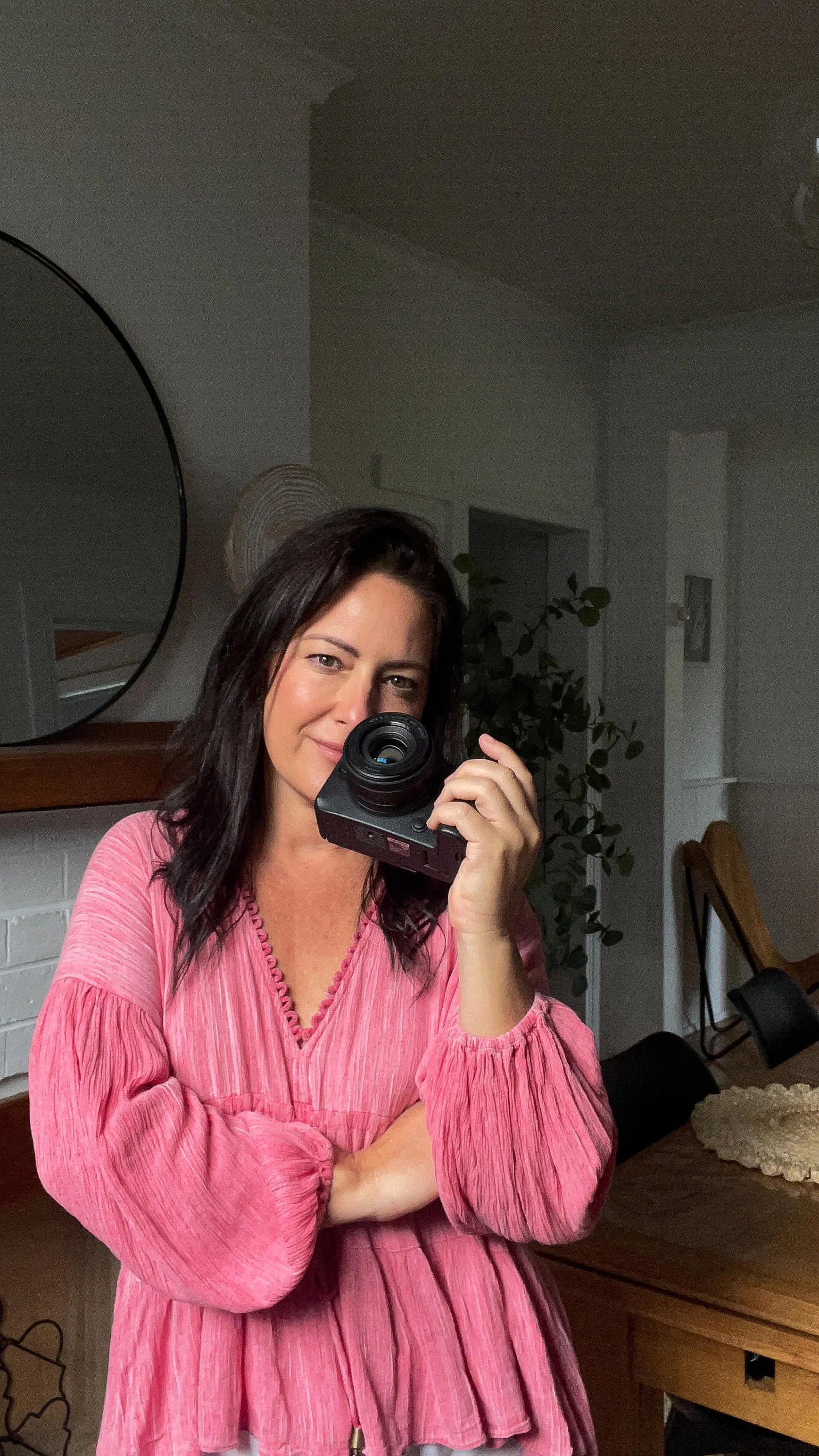 A woman taking a selfie in front of a mirror, wearing a pink blouse with long sleeves.