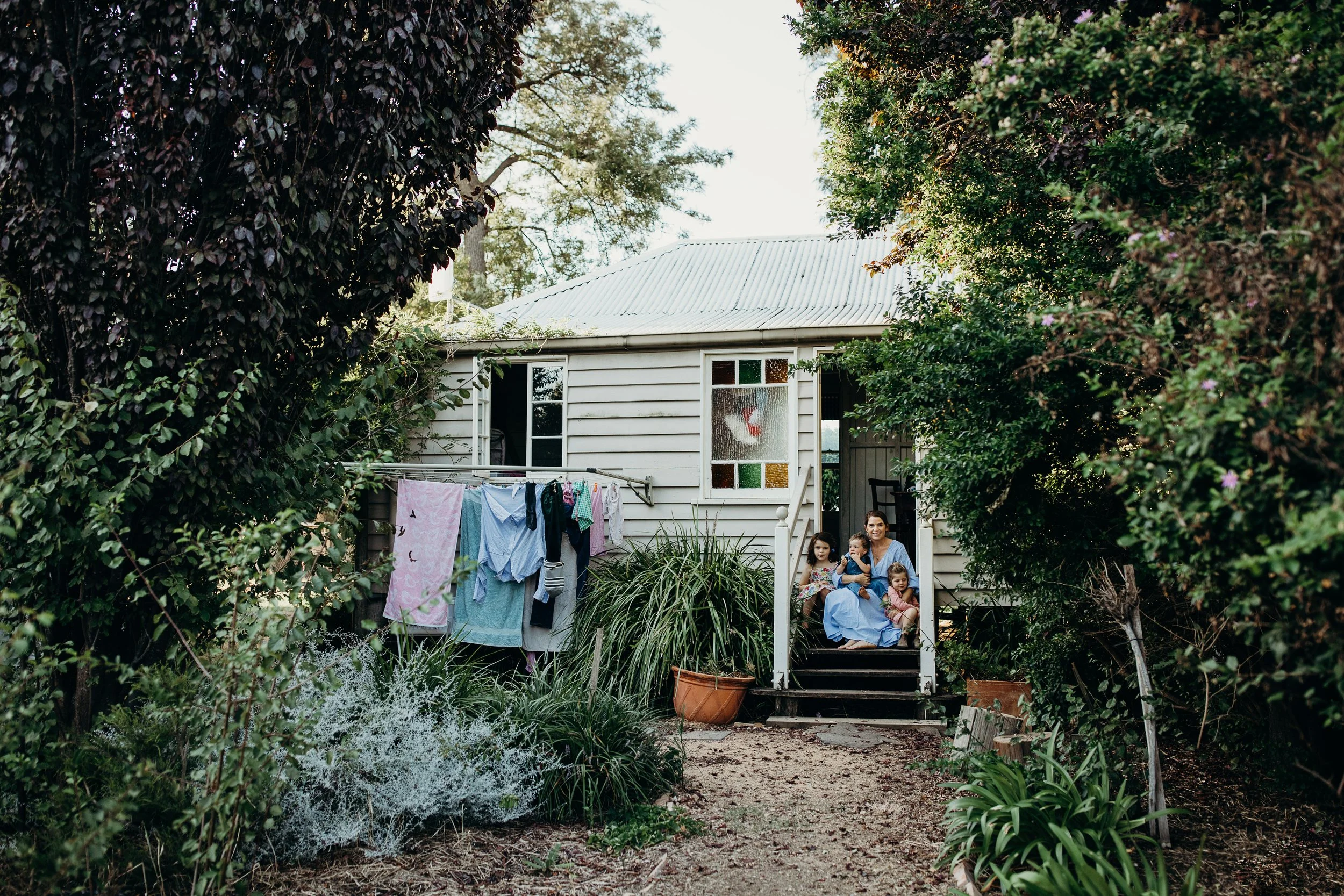 A woman and three children sitting on the front steps of a house surrounded by greenery with laundry hanging outdoors.