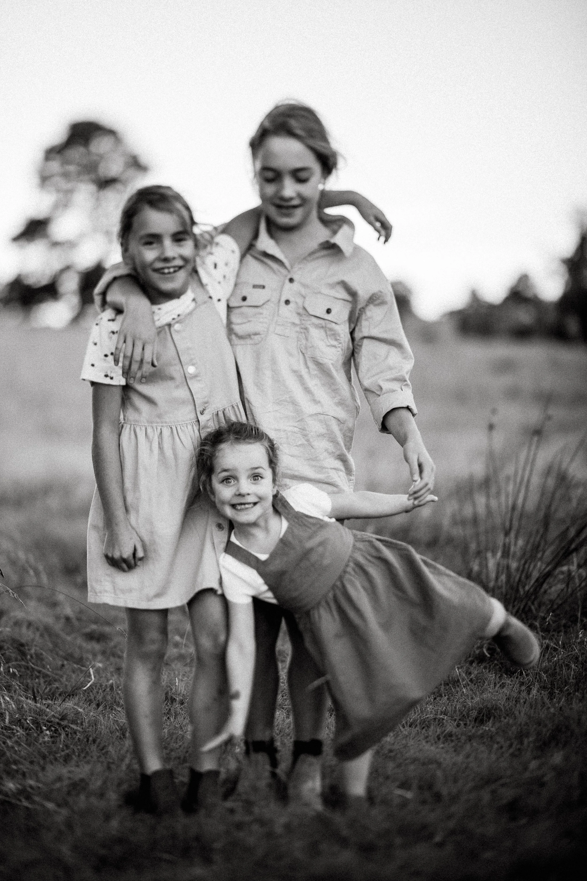 Three children outdoors, smiling and holding hands, with a blurred natural background, in black and white.