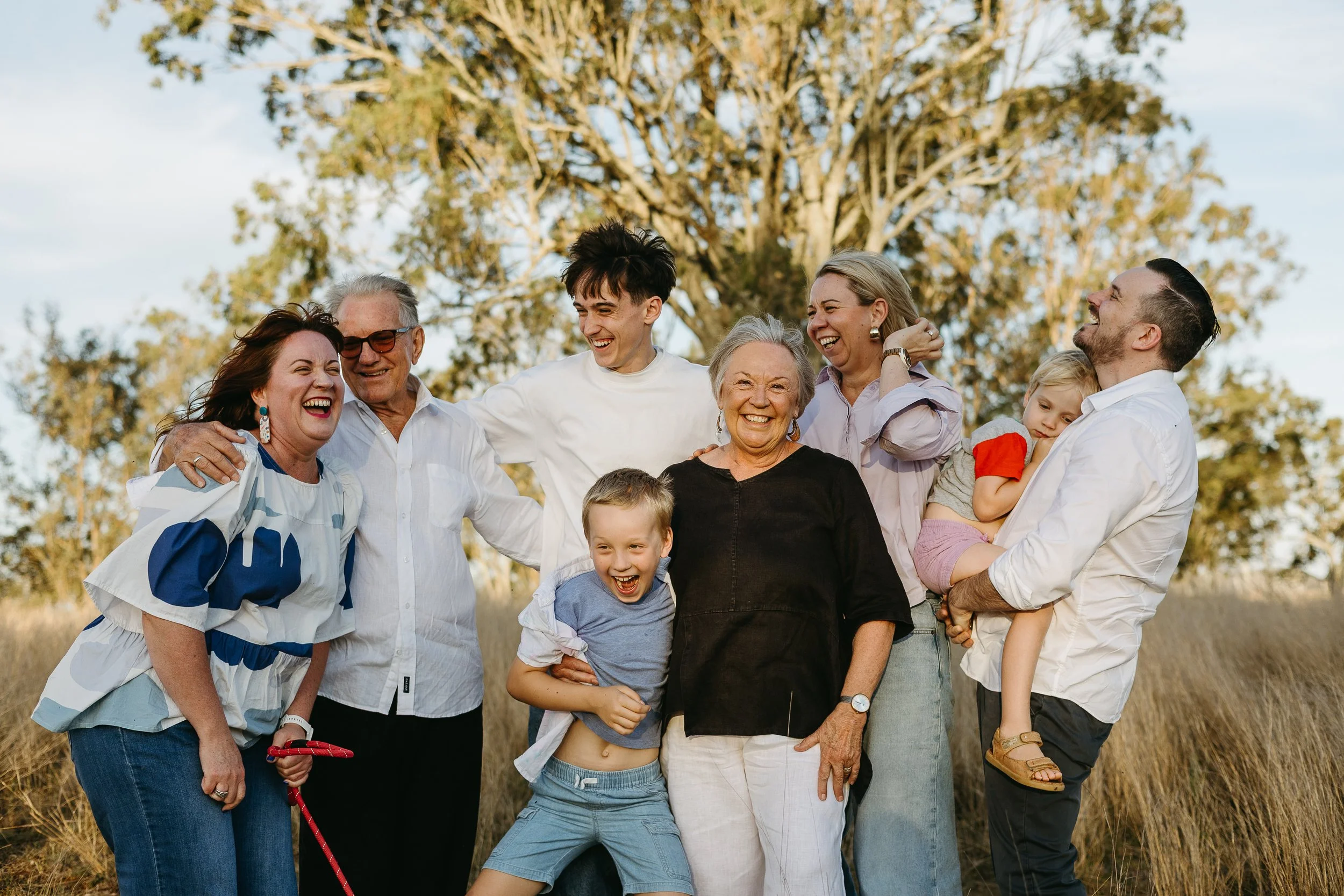 An extended family group laughing and smiling outdoors in a grassy area with trees in the background. Toowoomba and Brisbane Family Photo Packages and Prices