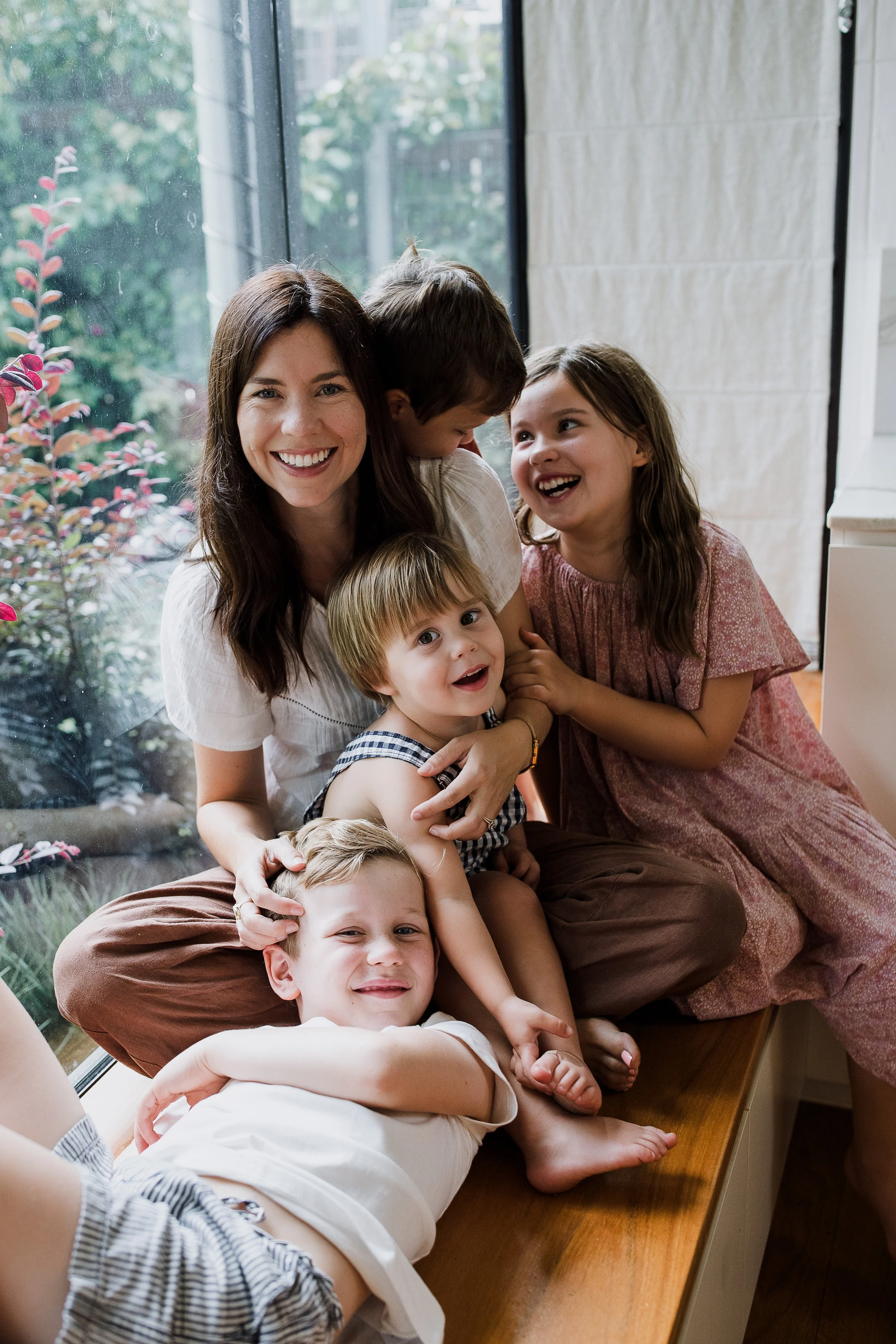 A woman and four children sitting on a window bench, smiling and playing together indoors, with a garden visible outside the window.
