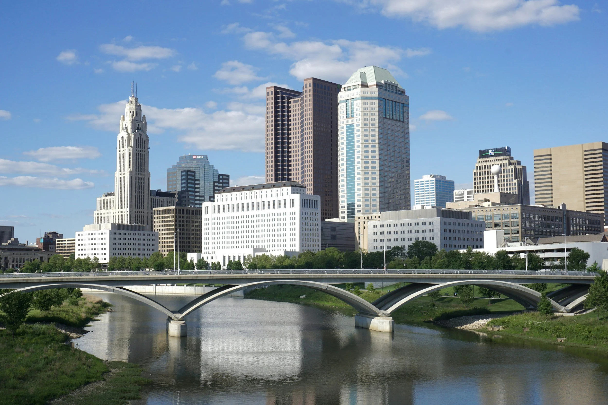 City skyline with tall skyscrapers and a river in the foreground, under a blue sky with scattered clouds.