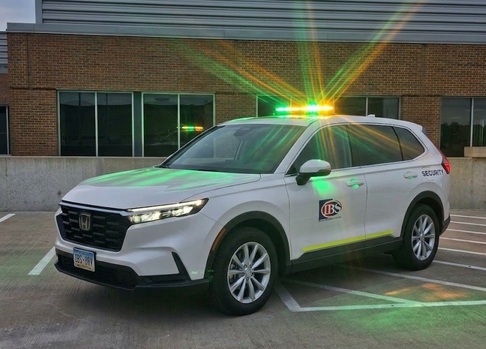 A white security SUV with flashing yellow and green lights parked in a lot near a brick building.