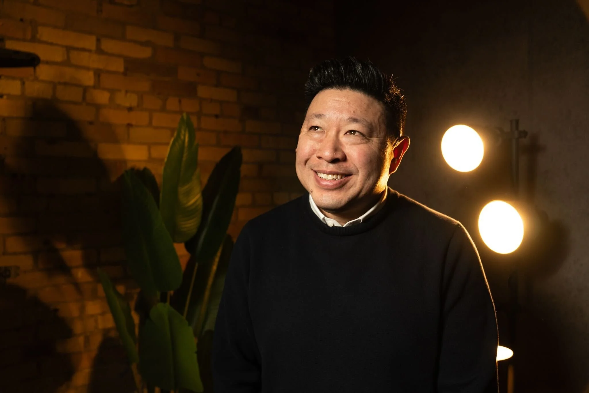 A man smiling, standing in a dimly lit room with a brick wall, green plants, and round lamps illuminating the background.