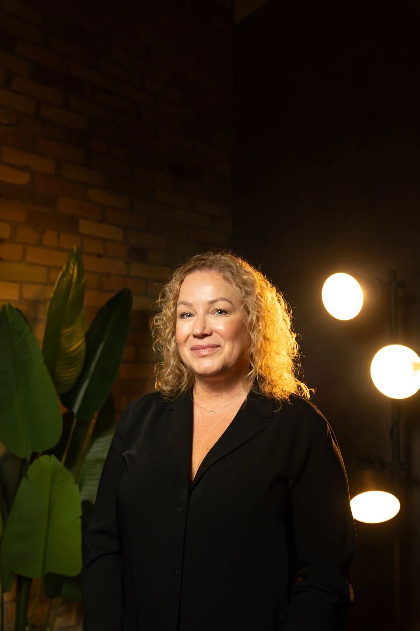 A woman with curly blonde hair, wearing a black blouse, standing in front of a brick wall with large green plant leaves on her left and bright round lights on her right.