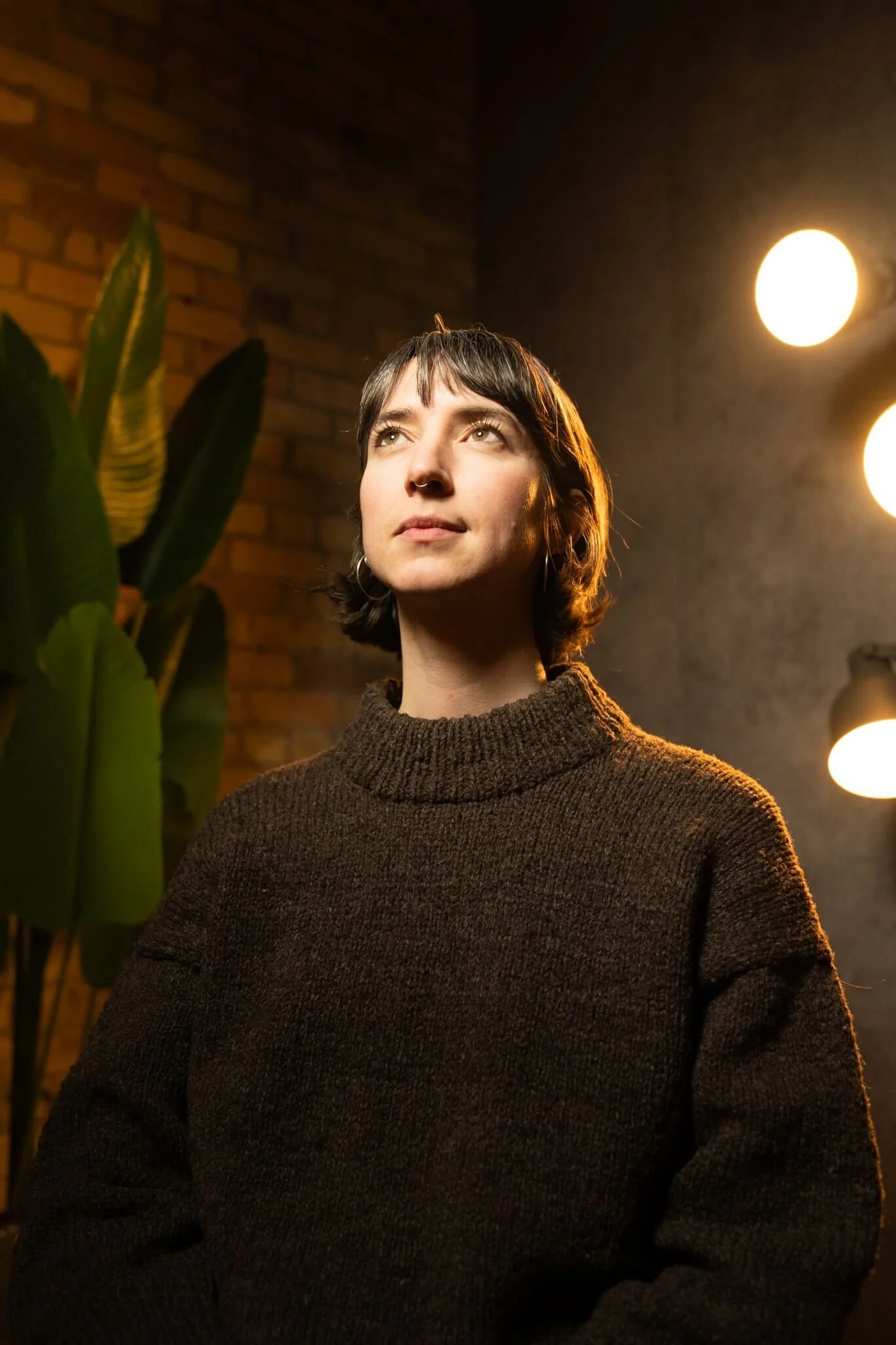 A young woman with short dark hair and earrings looking thoughtfully upward, standing indoors with a brick wall background, illuminated by warm lights.