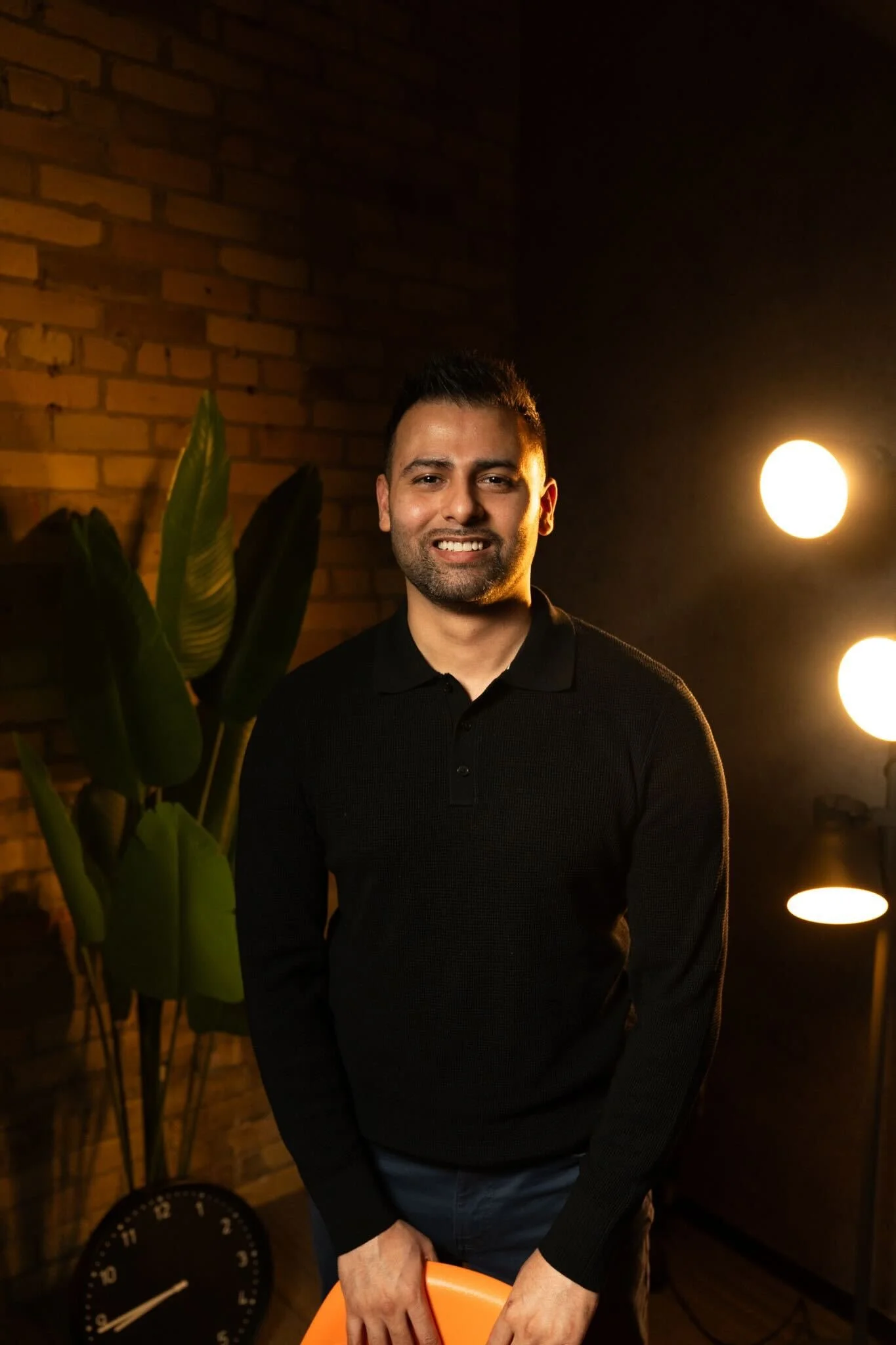 A smiling man with short dark hair and a beard, wearing a black sweater, standing indoors near a brick wall and a tall green plant. Two illuminated round lights are behind him on the right side of the image.