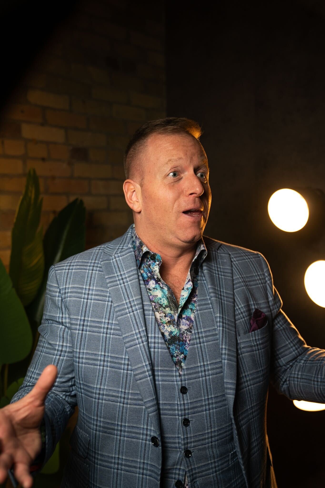A man with short hair and a surprised or expressive facial expression, wearing a plaid blazer and colorful shirt, standing in front of a brick wall and studio lights.