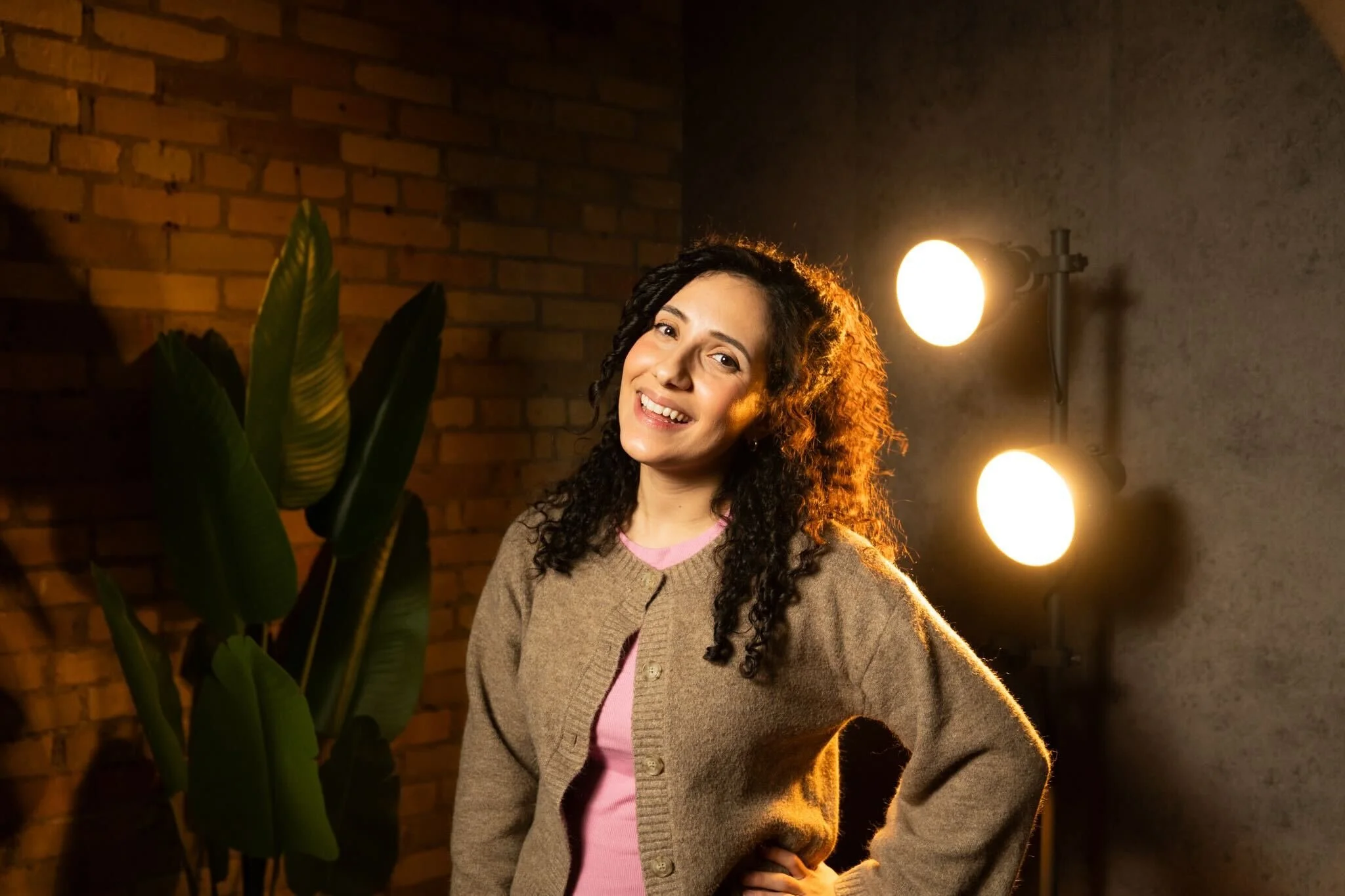 A woman with curly hair smiling at camera indoors with a brick wall and studio lights in the background.
