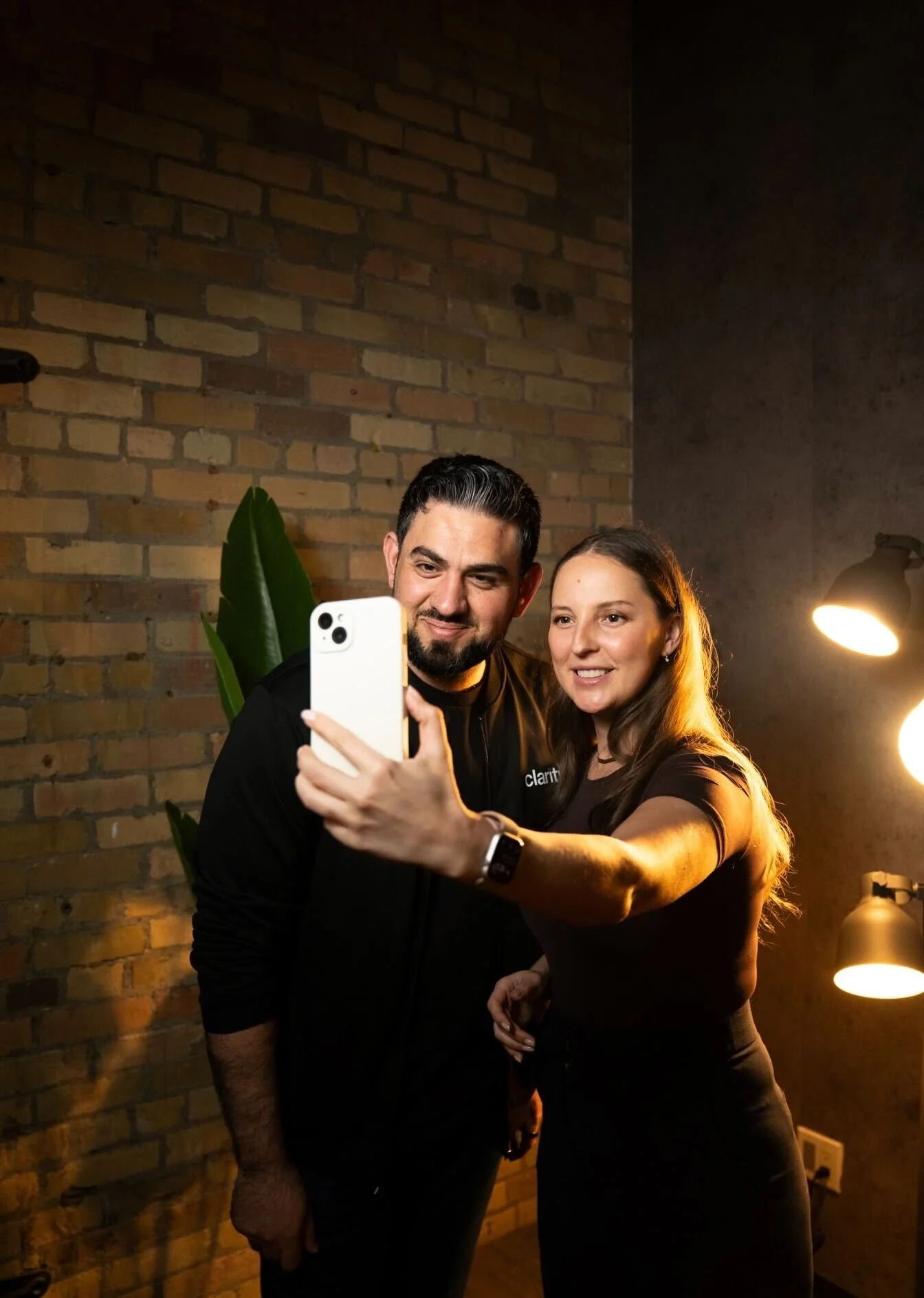 A man and woman taking a selfie together indoors with a brick wall background, warm lighting, and a large green plant behind them.