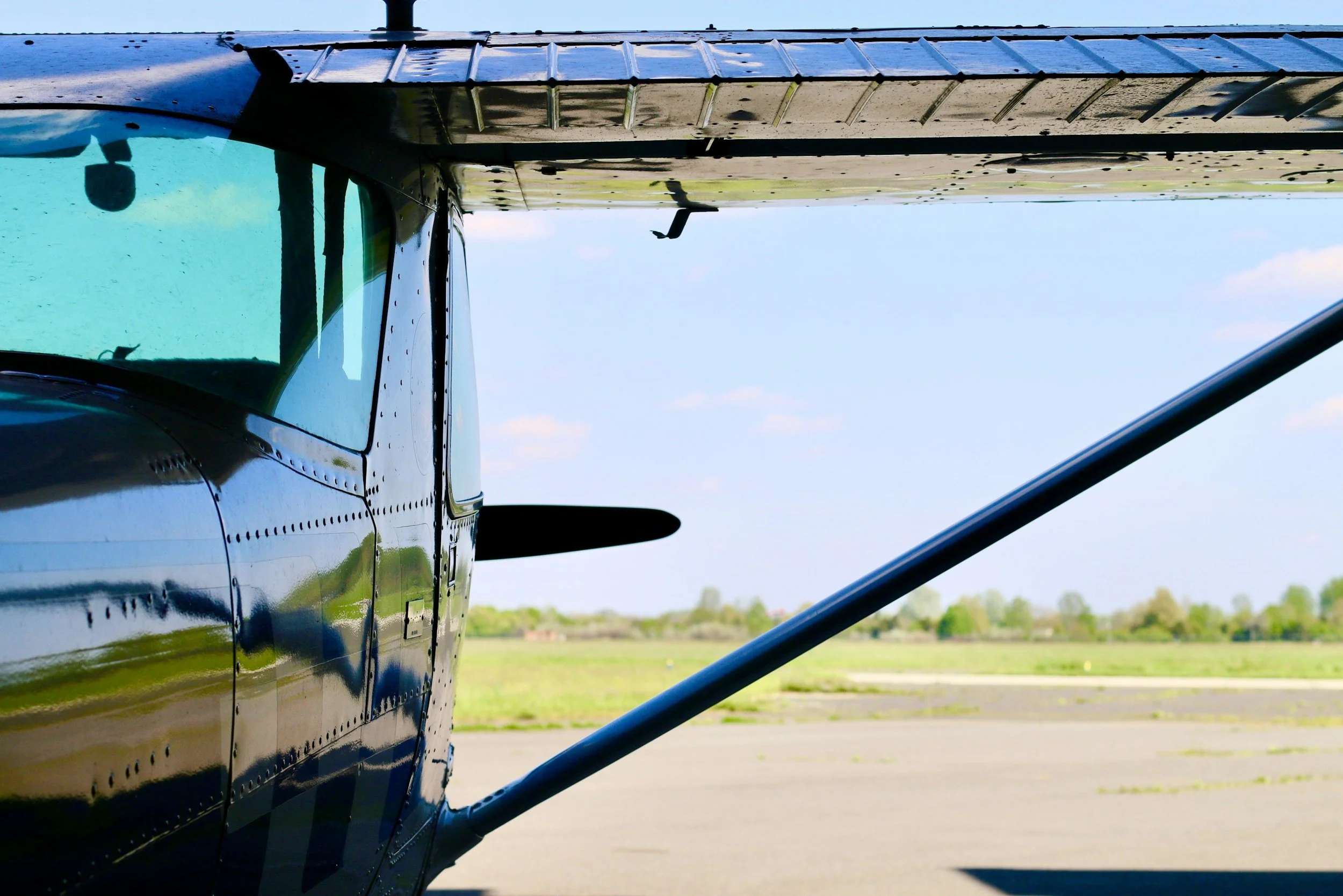 Close-up of a small aircraft on a runway, showing the cockpit, wing, and tail against a clear sky and green landscape.