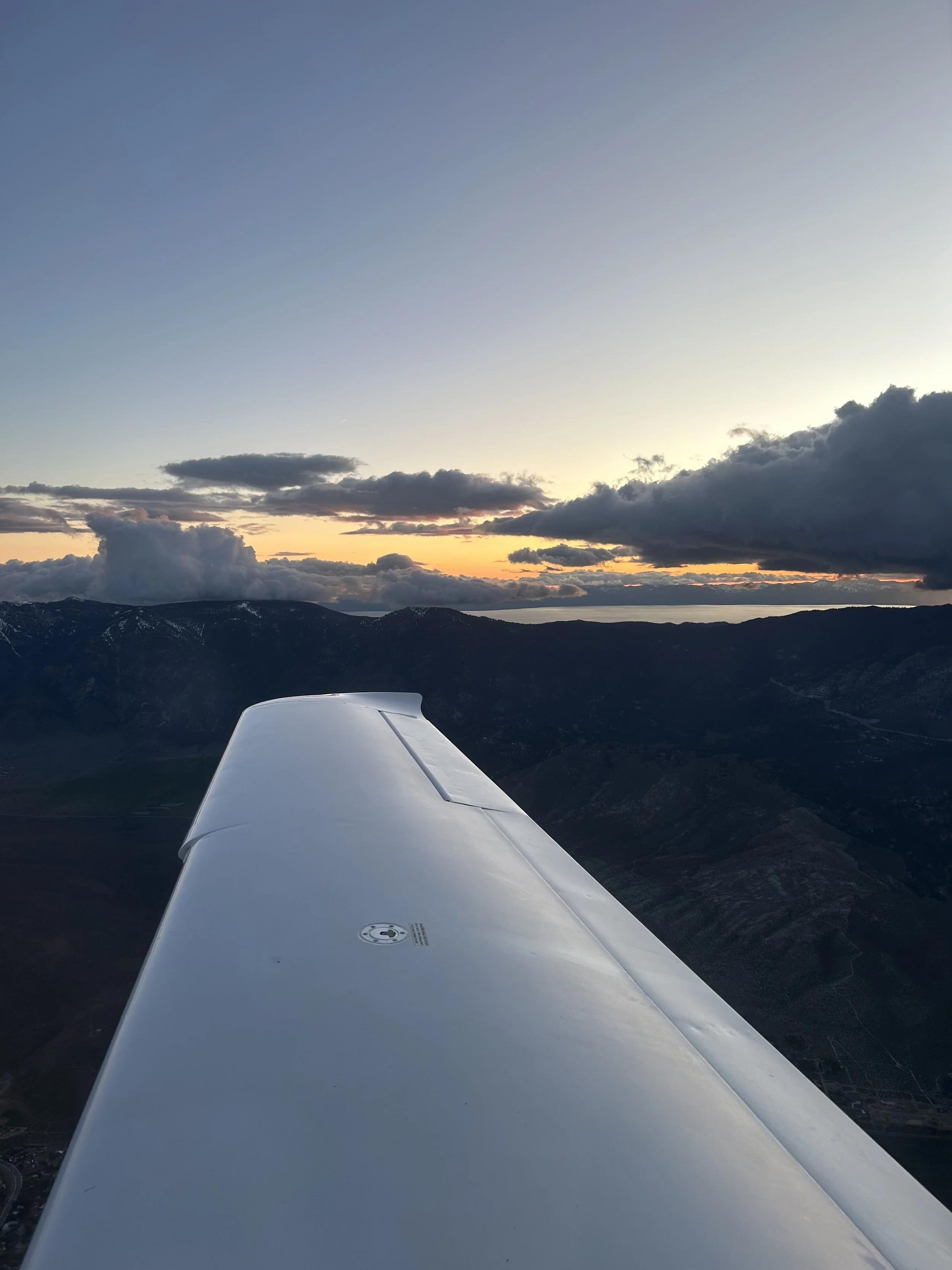 A view from an airplane wing during sunset over mountains and clouds.