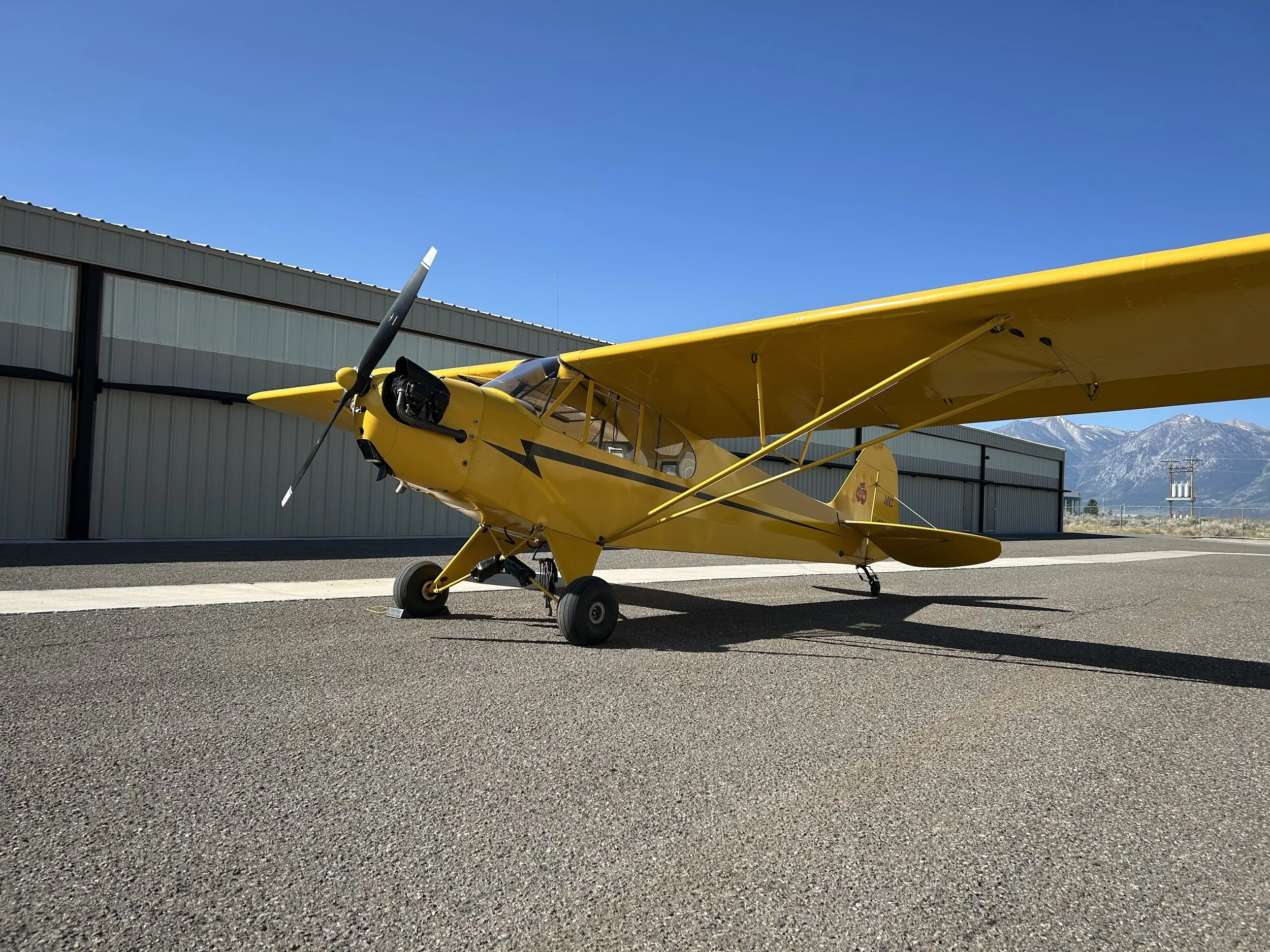 A bright yellow small aircraft parked on a tarmac in front of a hangar with mountains in the background and a clear blue sky.