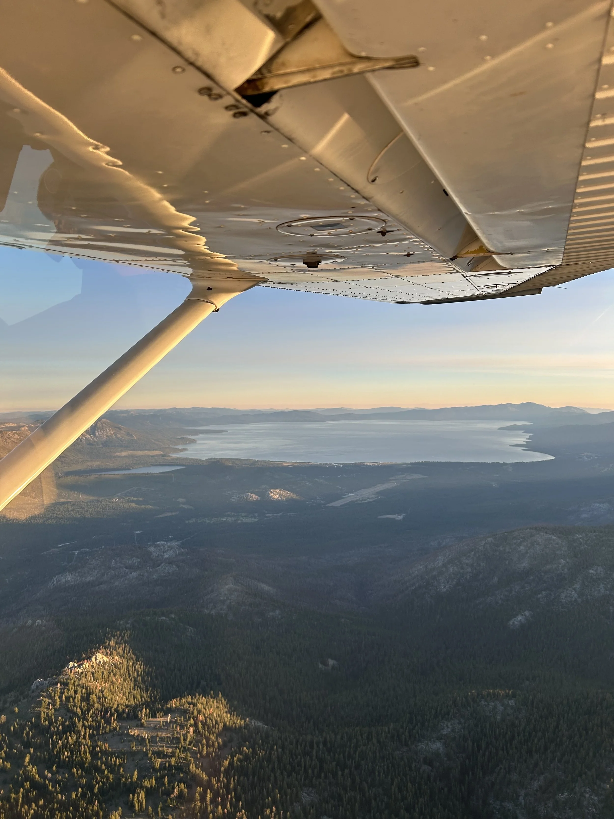 Aerial view from an airplane showing a landscape with a large lake, mountains, and forested land during sunset.