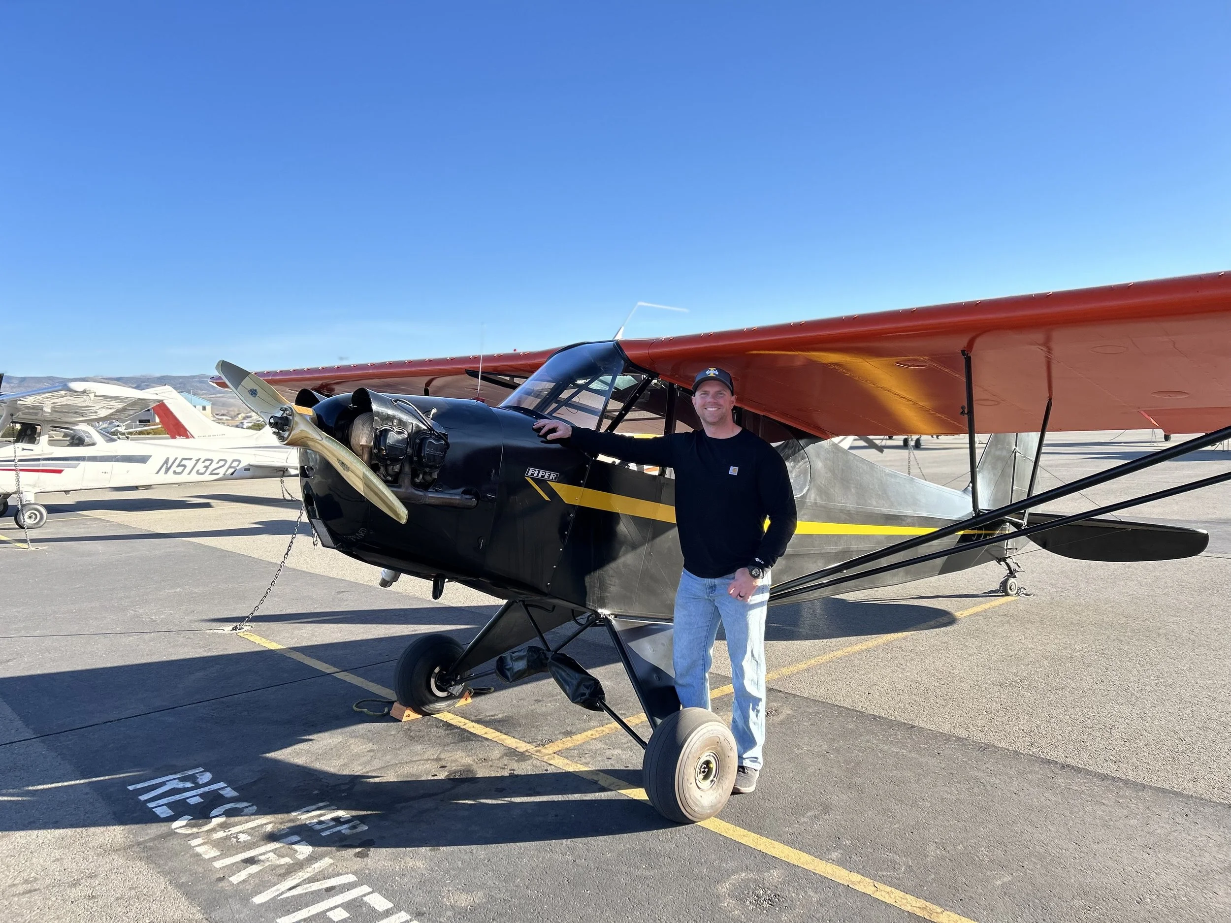 Man standing next to a small black and red propeller airplane on an airport tarmac, with a white airplane in the background under a clear blue sky.
