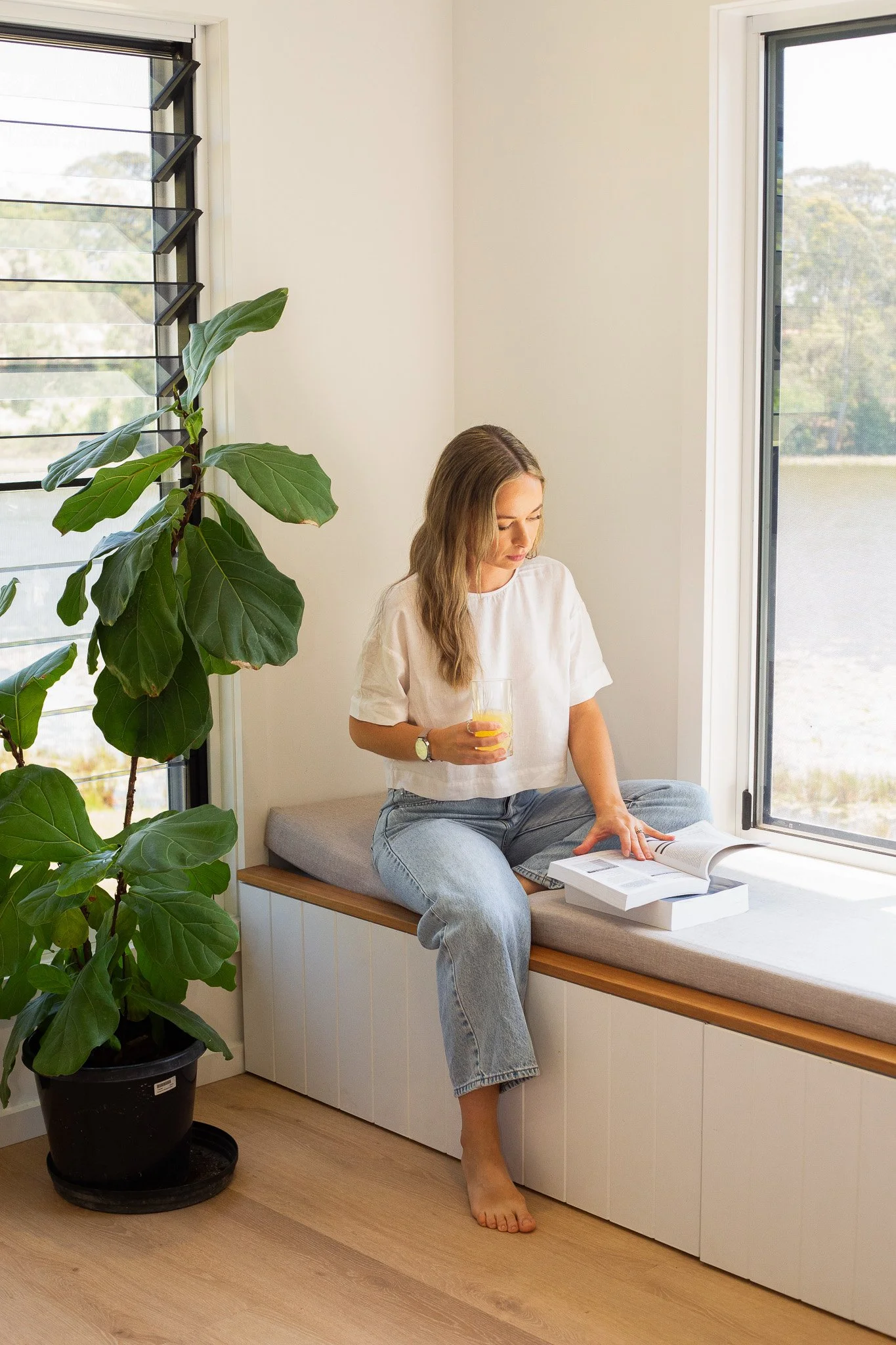 A woman sitting by a large window, reading a book, with a glass of orange juice in her hand, inside a bright room with a potted plant nearby.