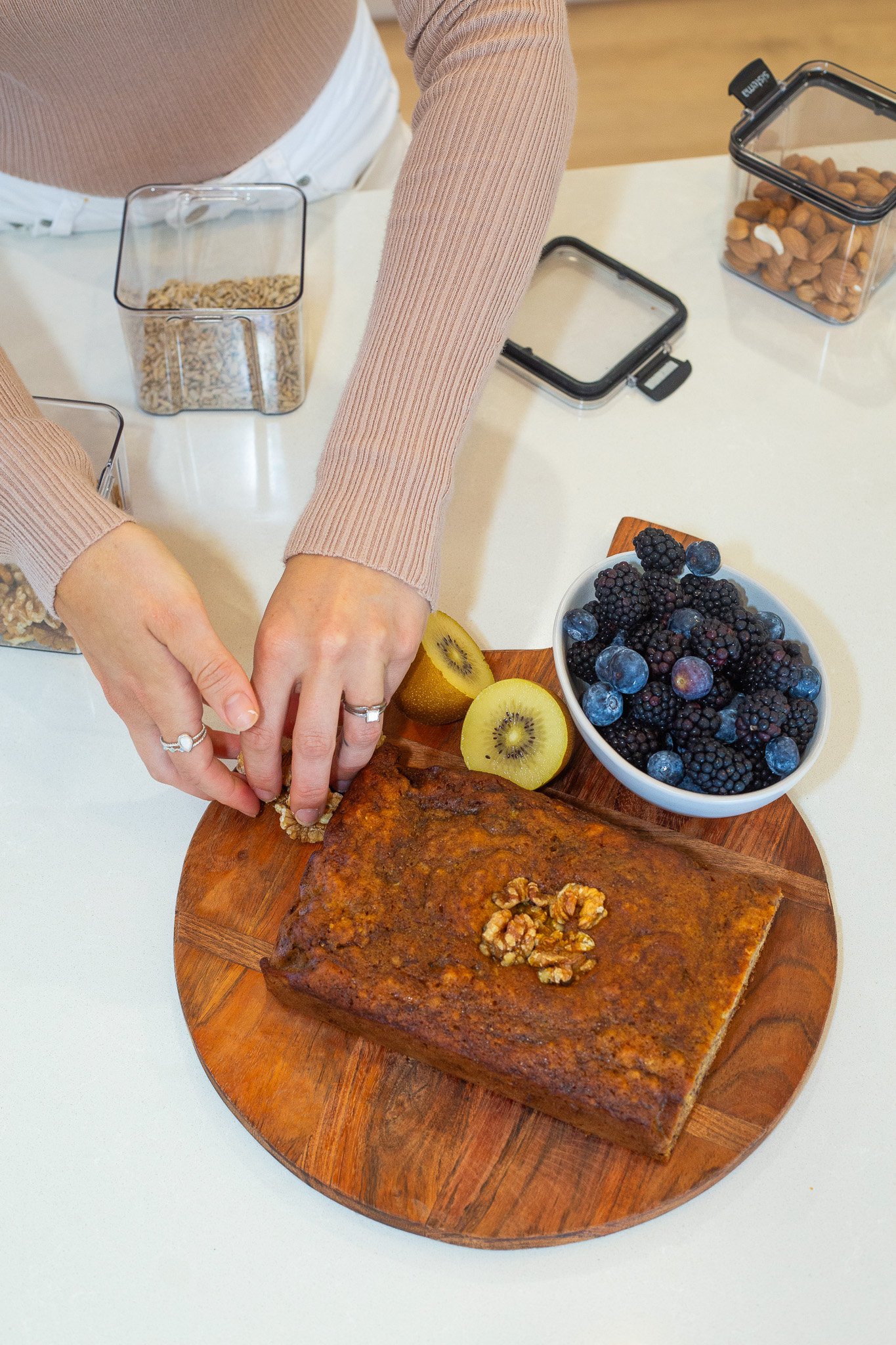 Person placing walnuts on a loaf cake on a wooden serving board, with a bowl of mixed berries and sliced kiwi on the board. There are food containers and a phone case on the table.