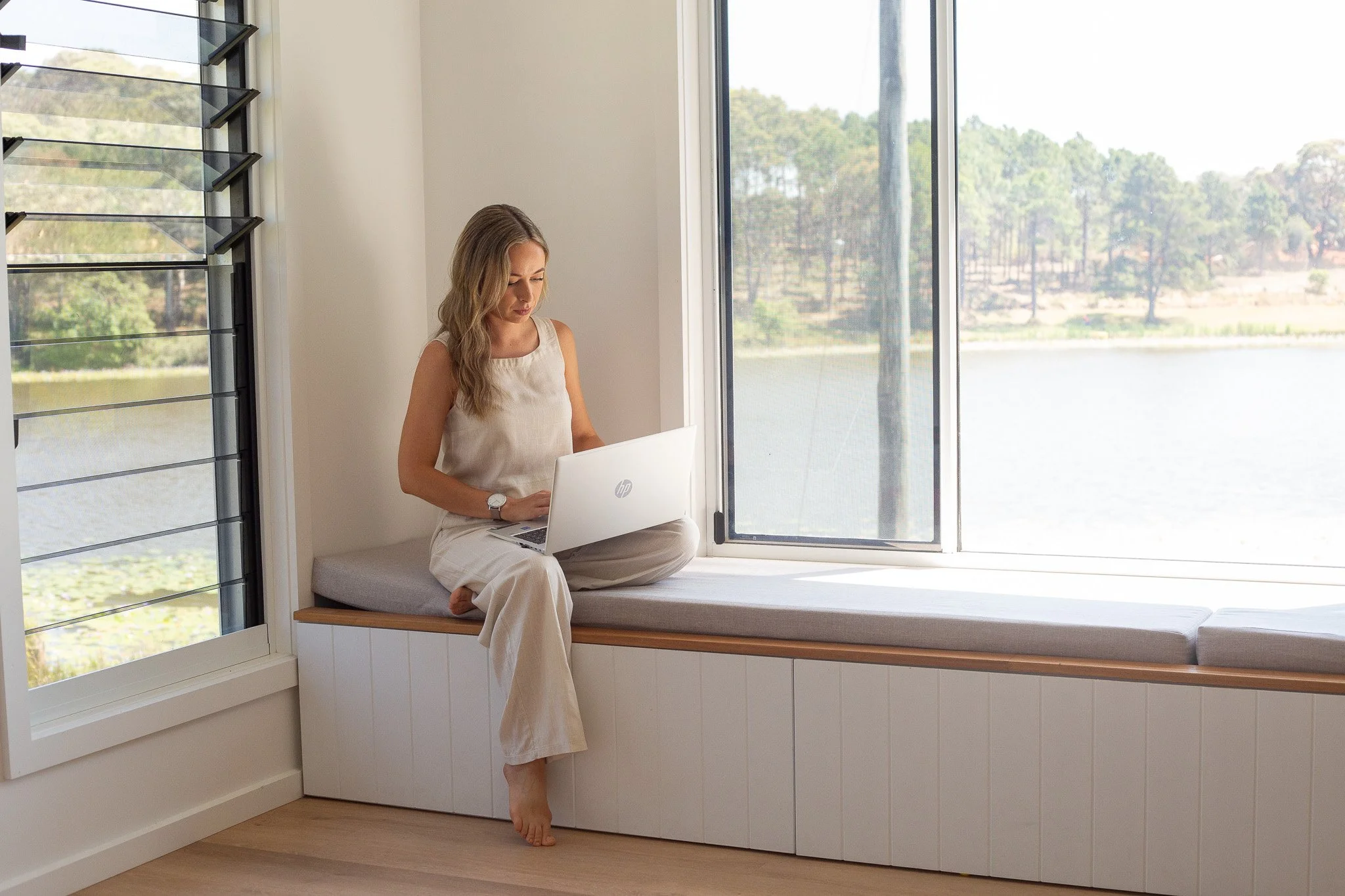 Woman sitting on a window bench using a laptop with a lake and trees outside the window.