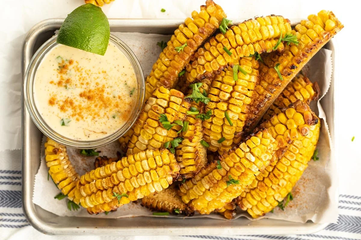 Baked corn on the cob with chopped cilantro, served with a side of dipping sauce garnished with a lime wedge.