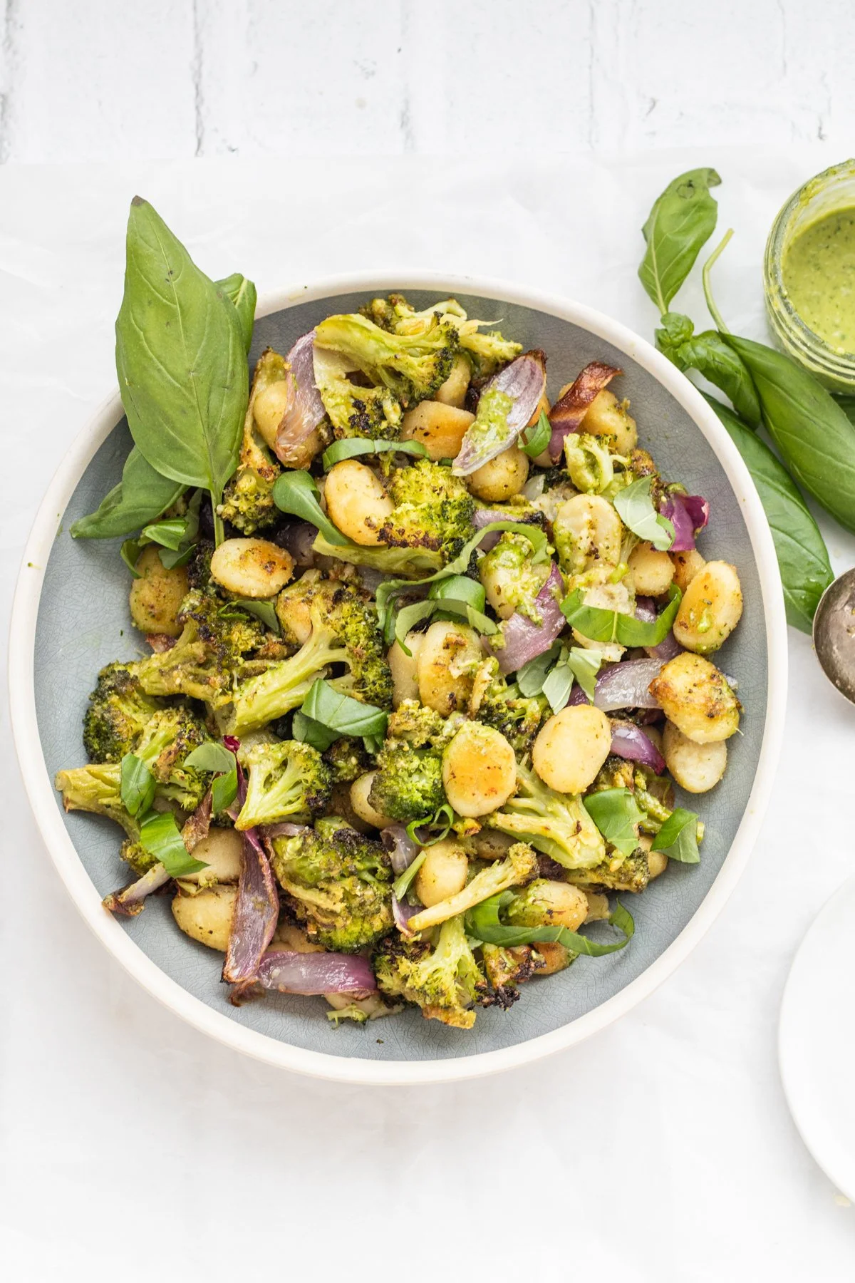 A bowl of roasted broccoli, cauliflower, chickpeas, and red onions garnished with fresh basil and green herbs, served on a white surface.