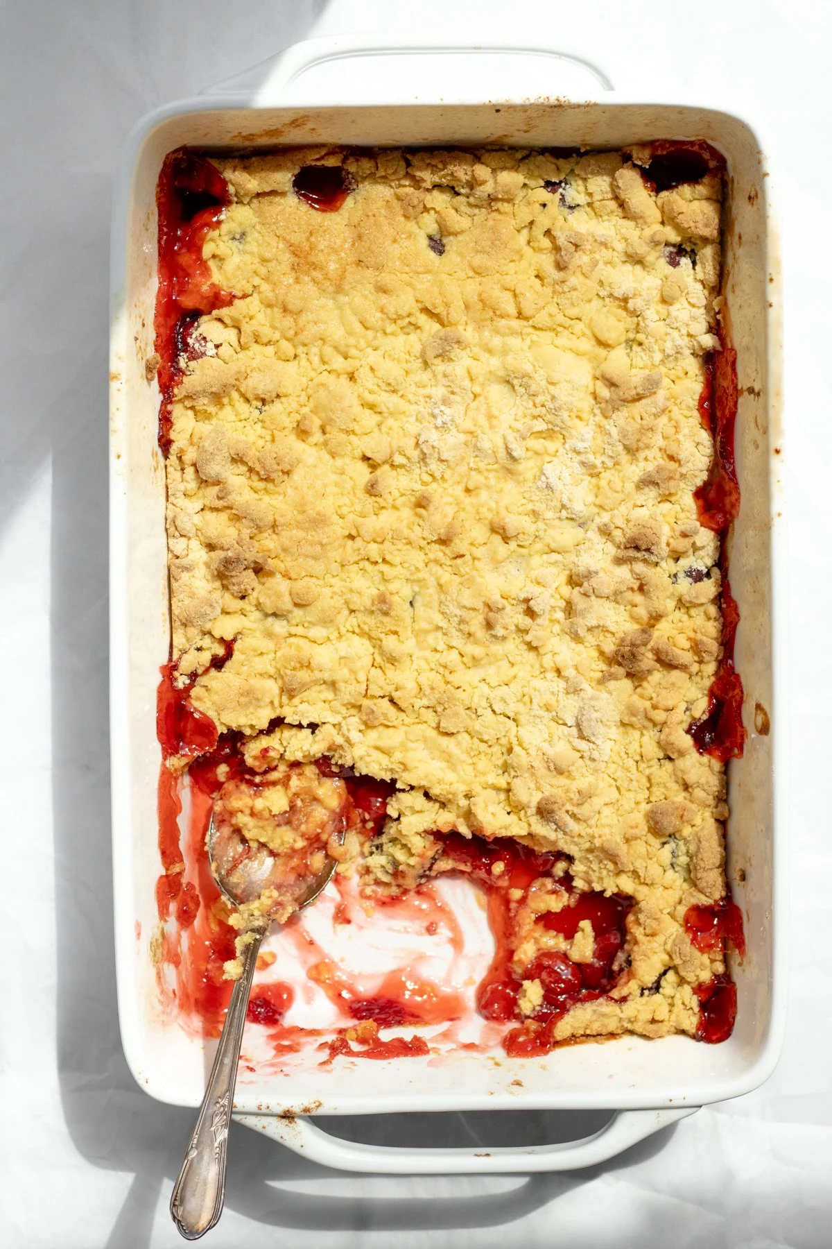 A partially eaten cherry crumble dessert in a white rectangular baking dish, with a spoon resting inside.