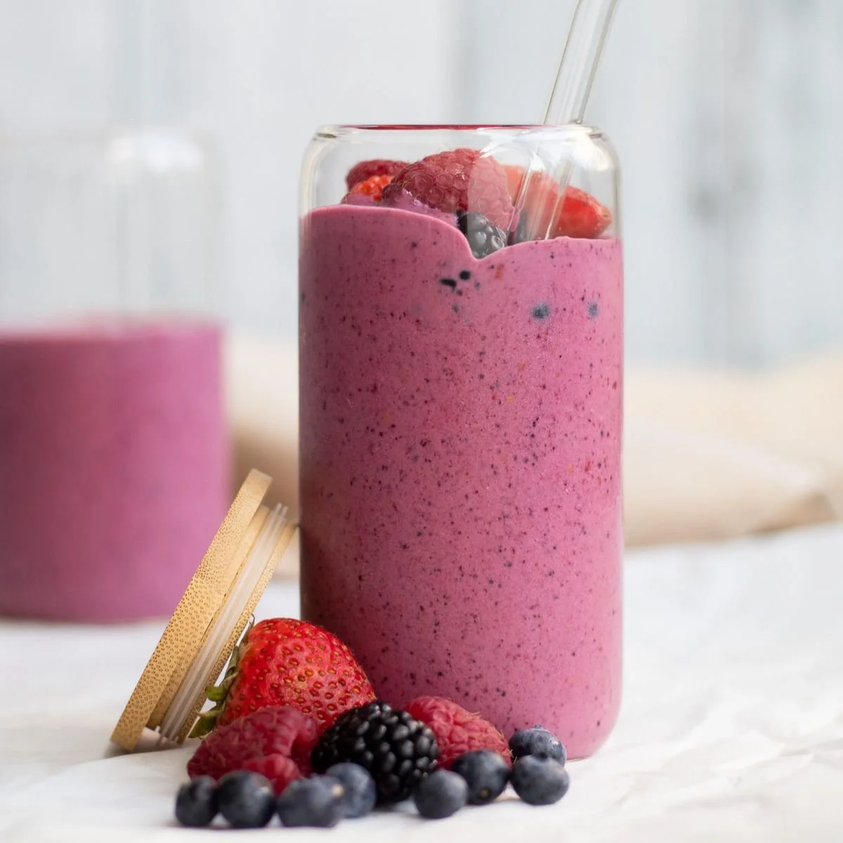 A tall glass filled with a pinkberry smoothie topped with mixed berries, with a straw, on a white surface, with fresh berries and a wafer cookie beside it.