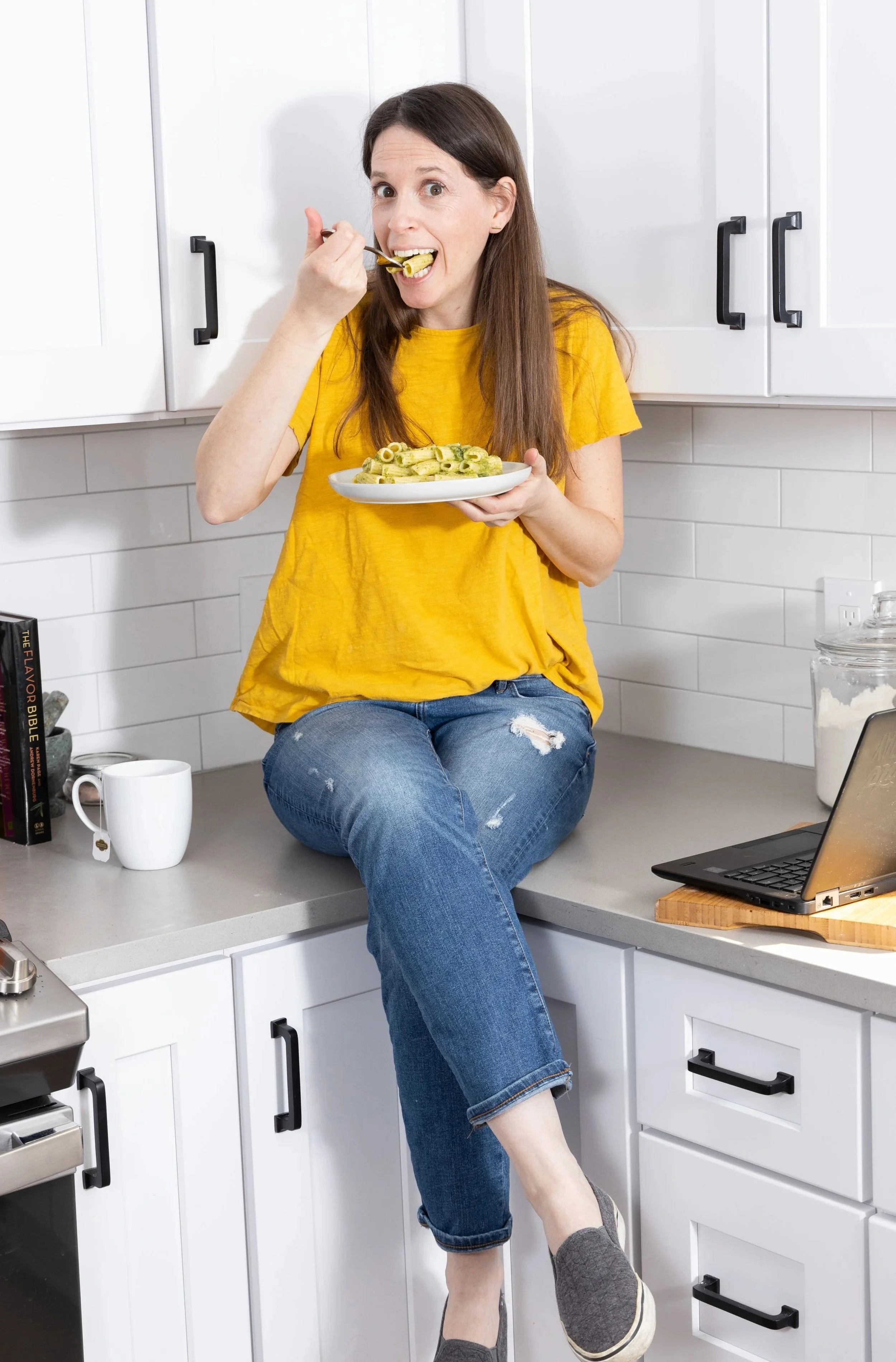 A woman in a yellow shirt and ripped jeans sitting on a kitchen countertop, eating pasta with a fork, holding a plate of pasta, in a white kitchen with black handles on cabinets.