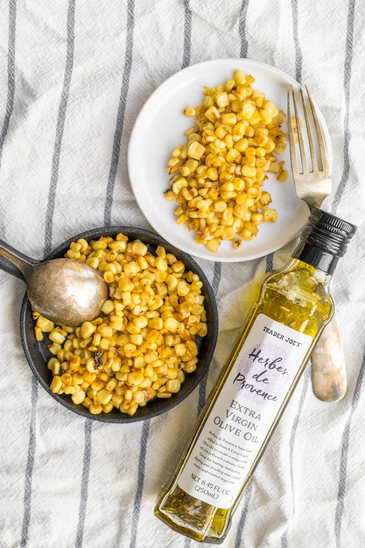 Cooked yellow corn in a small skillet and on a white plate with a fork, a bottle of extra virgin olive oil, and a cloth with gray stripes underneath.
