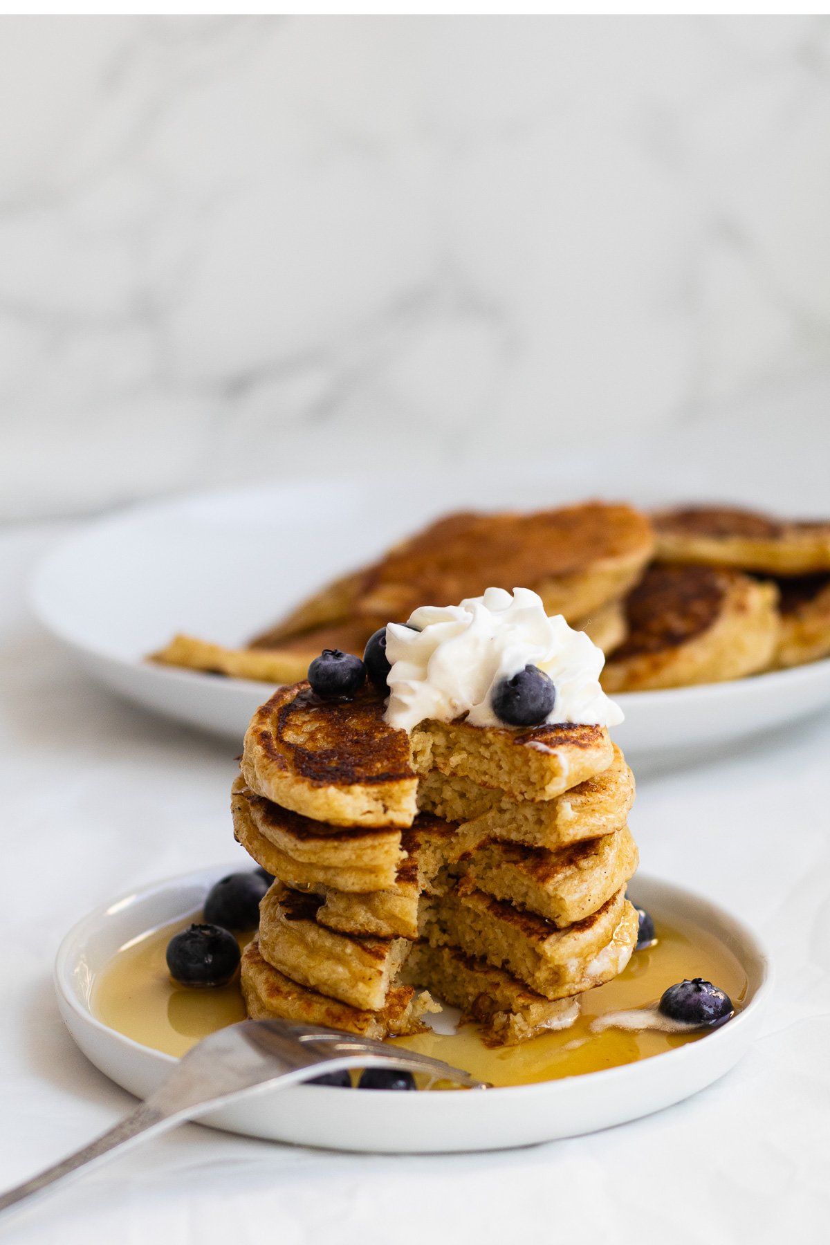 Stack of blueberry pancakes topped with whipped cream and blueberries, served with syrup on a white plate, with more pancakes in the background.
