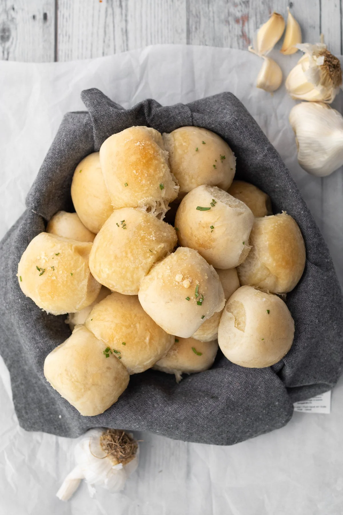 Basket of freshly baked bread rolls garnished with chopped chives on a gray cloth, with garlic cloves nearby on a white wooden surface.