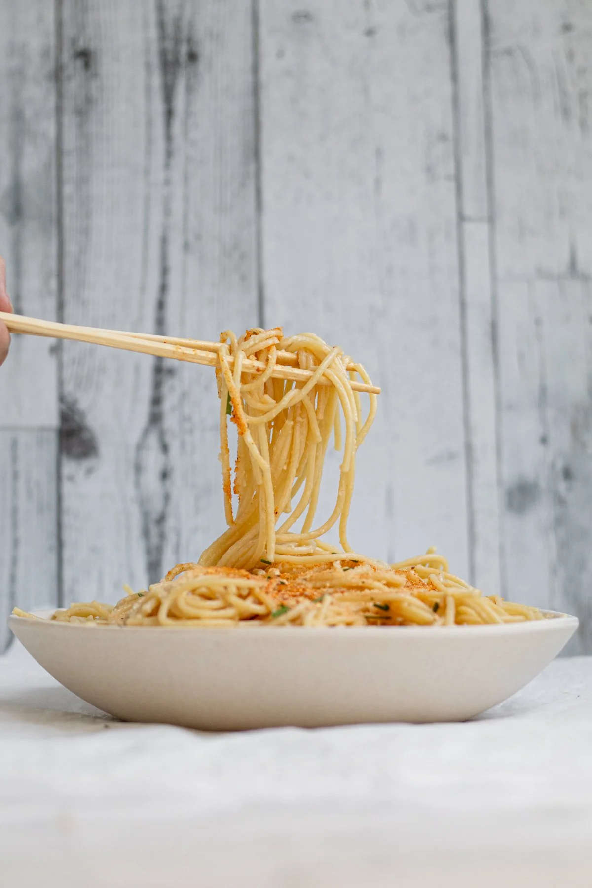 Close-up of a hand holding chopsticks lifting a portion of spaghetti from a bowl of pasta on a white surface against a wooden background.