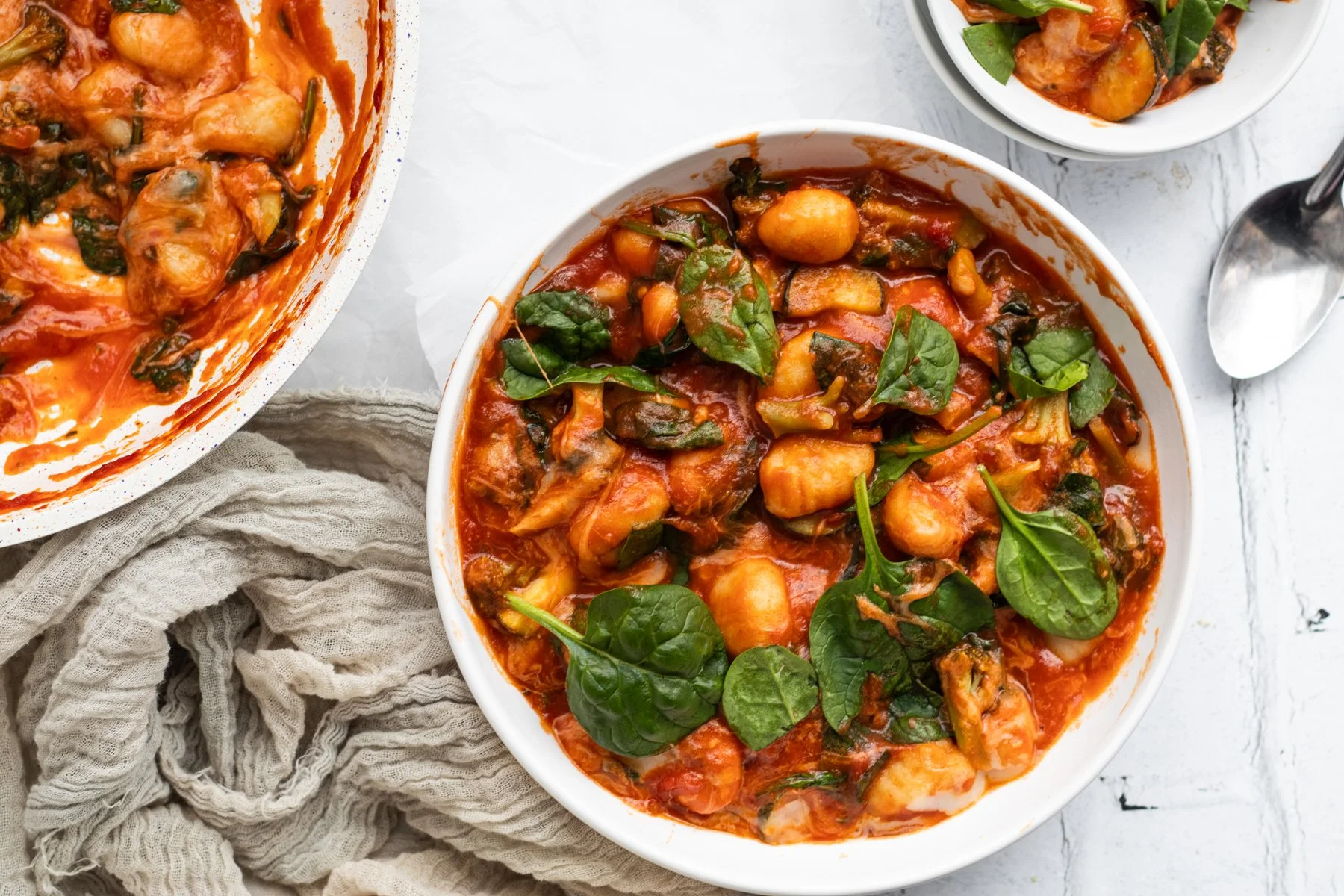 A bowl of gnocchi with tomato sauce and fresh spinach leaves, accompanied by a smaller bowl of the same dish and a metal spoon on a white marble surface.