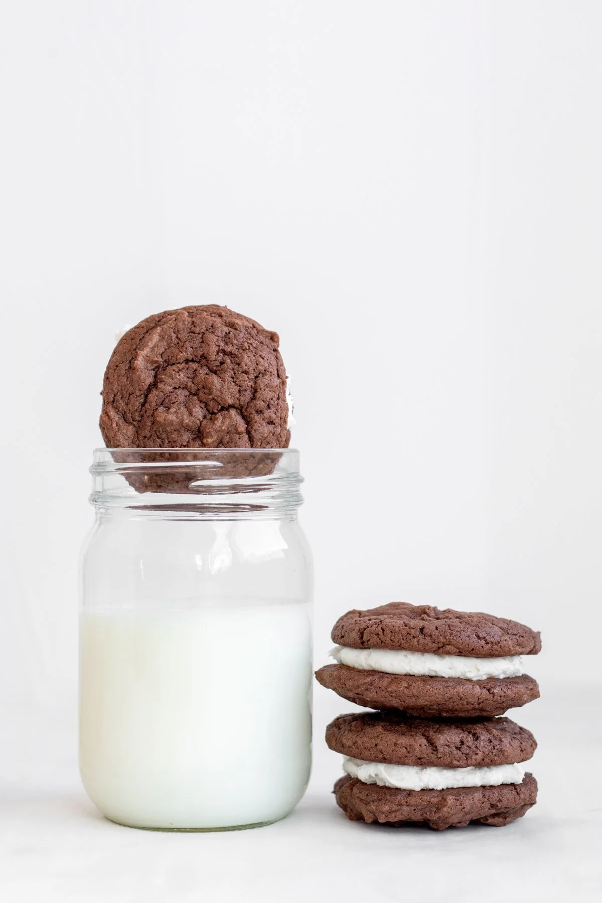 Chocolate cookies and cookie ice cream sandwiches beside a jar of milk on a white background.