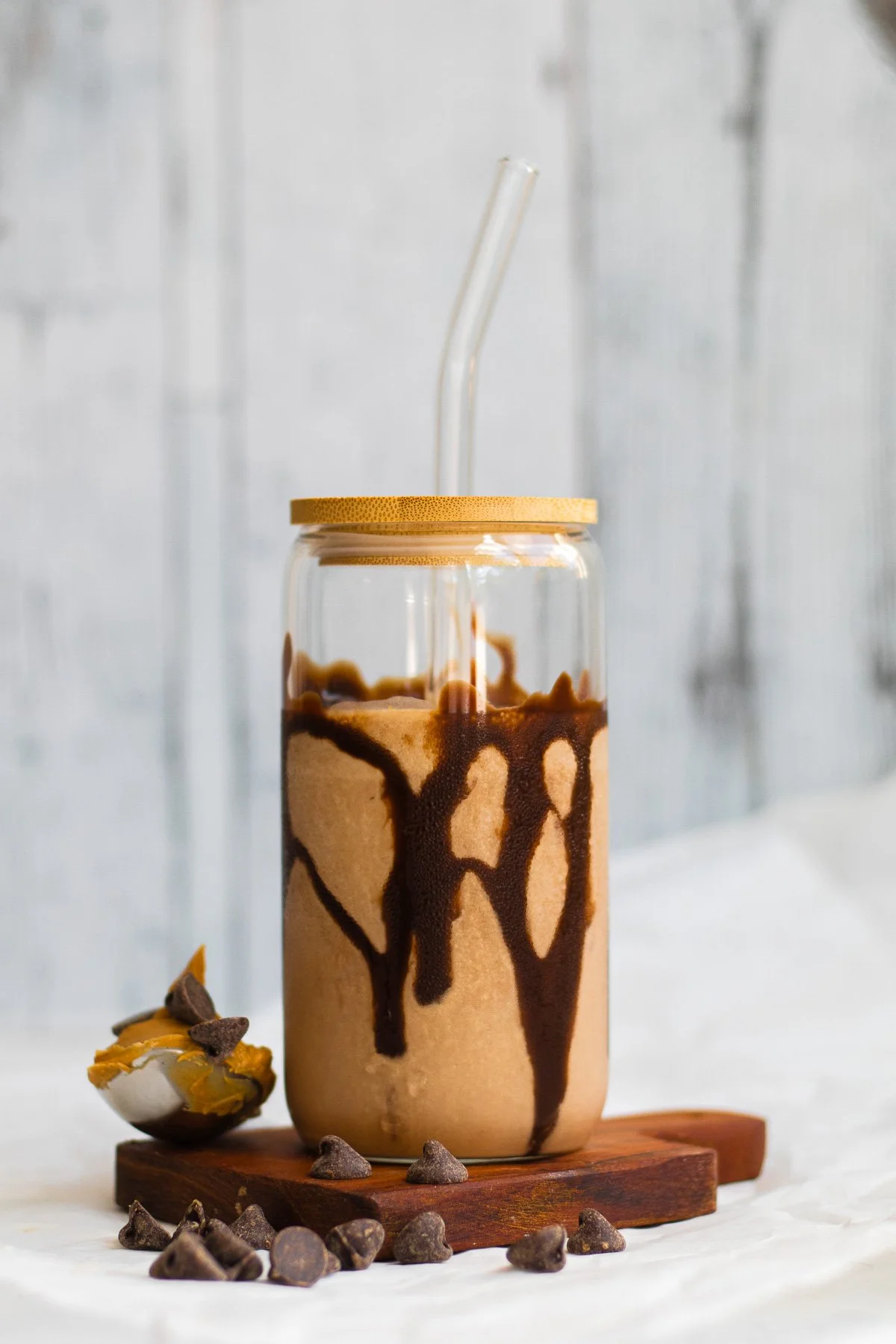 Chocolate milkshake in a glass jar with a bamboo lid, topped with chocolate syrup. The jar has a bendy straw and is placed on a wooden board with chocolate chips and a partially eaten cookie beside it.