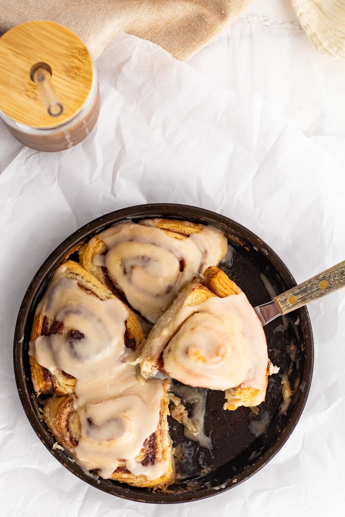 A baking dish with cinnamon rolls topped with icing, partly eaten, with a fork resting inside. A container of cinnamon and a jar of icing are visible in the background.