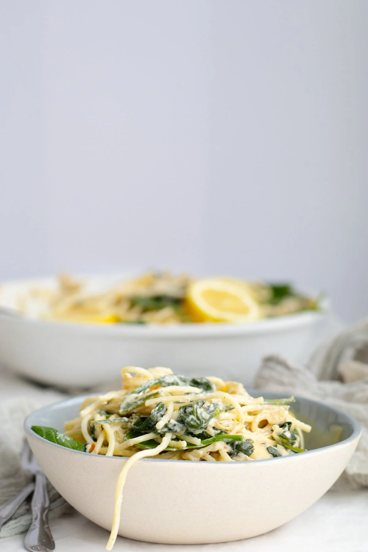 A bowl of creamy pasta with spinach on a white surface, with more pasta and lemon slices in a dish in the background.