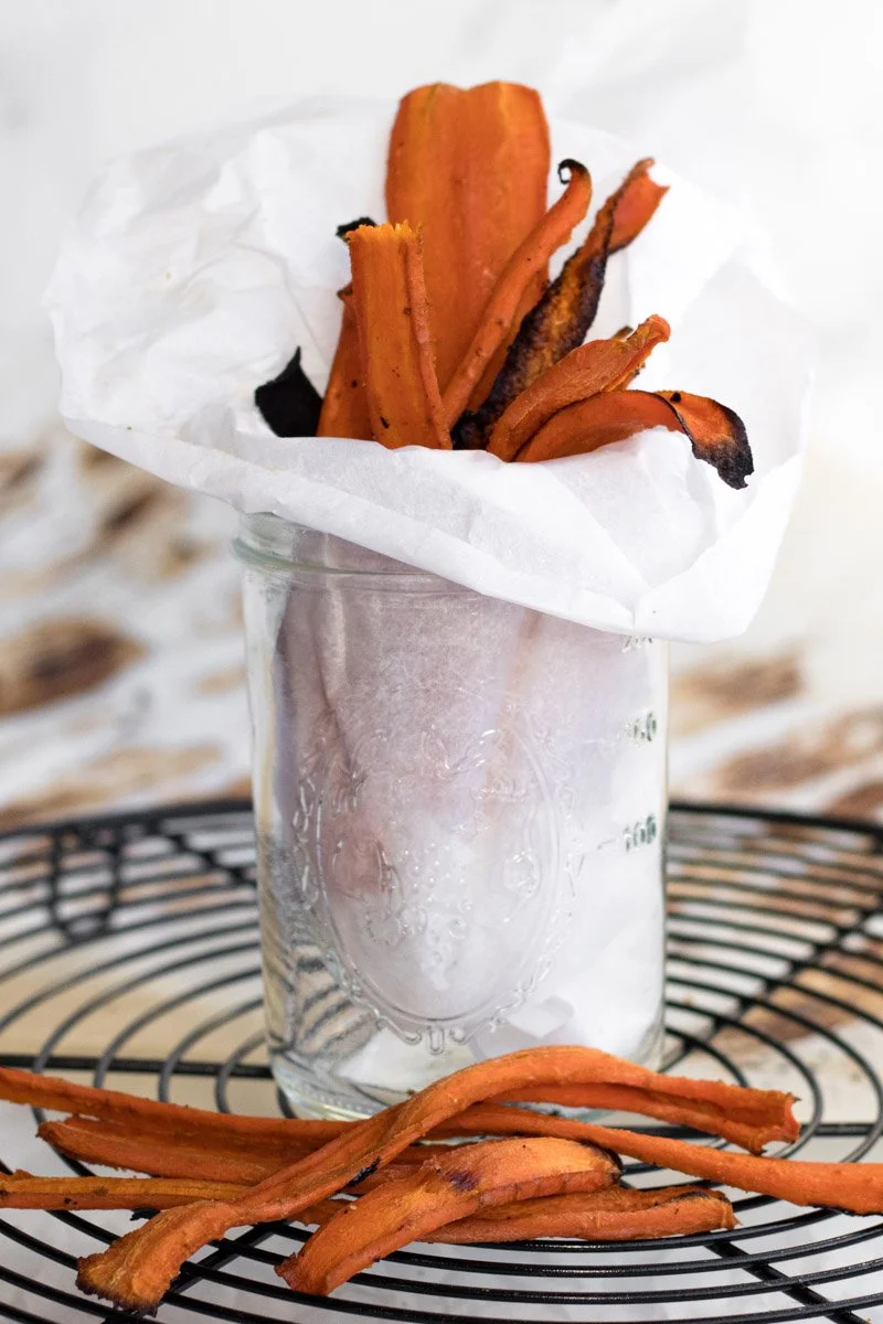 Sweet potato peels inside a glass jar lined with white paper, with some peels scattered on a wire cooling rack.
