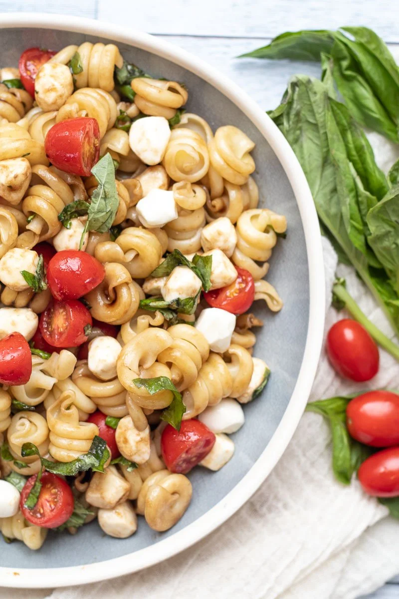 Pasta salad with corkscrew pasta, cherry tomatoes, mozzarella balls, basil, and grilled chicken in a white bowl with fresh basil leaves outside.