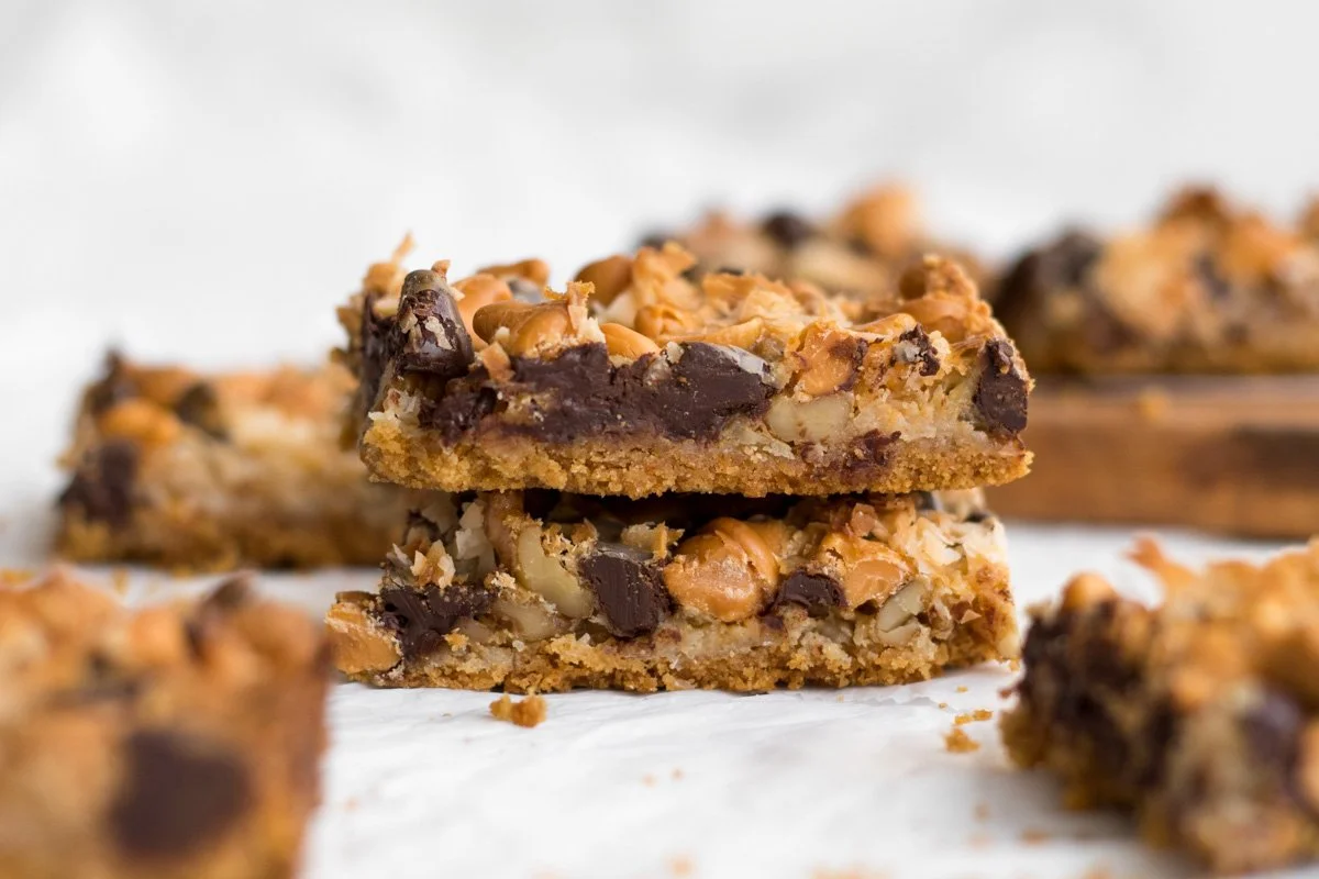 Close-up of homemade chocolate chip and peanut butter granola bars on white parchment paper, with more bars and a wooden tray in the background.