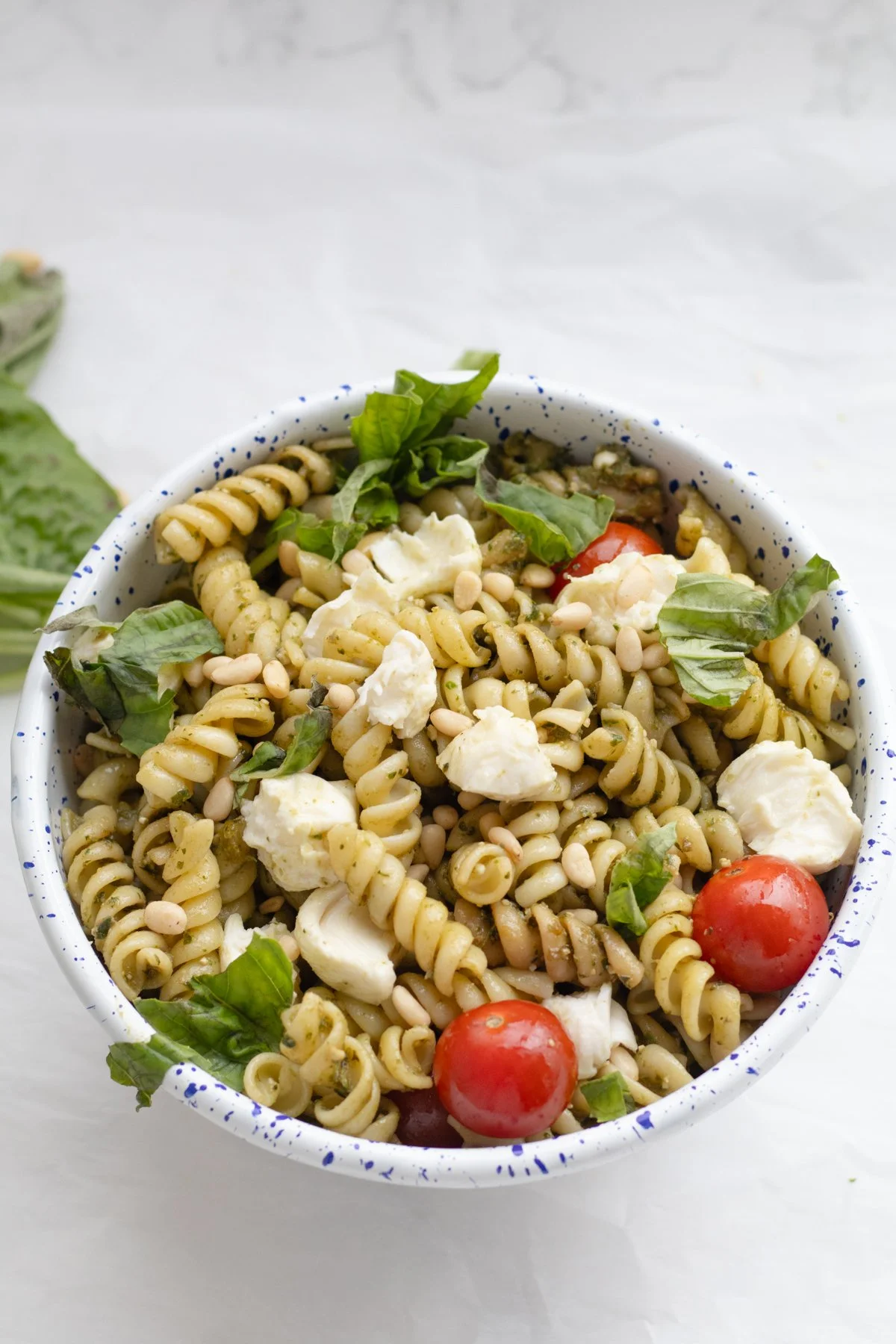 A bowl of pasta salad with cherry tomatoes, mozzarella cheese, basil leaves, and pine nuts on a white surface.