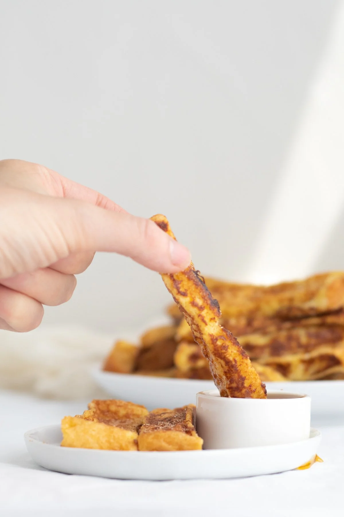Hand dipping crispy bacon into a small white bowl of syrup, with a plate of bacon strips in the background.