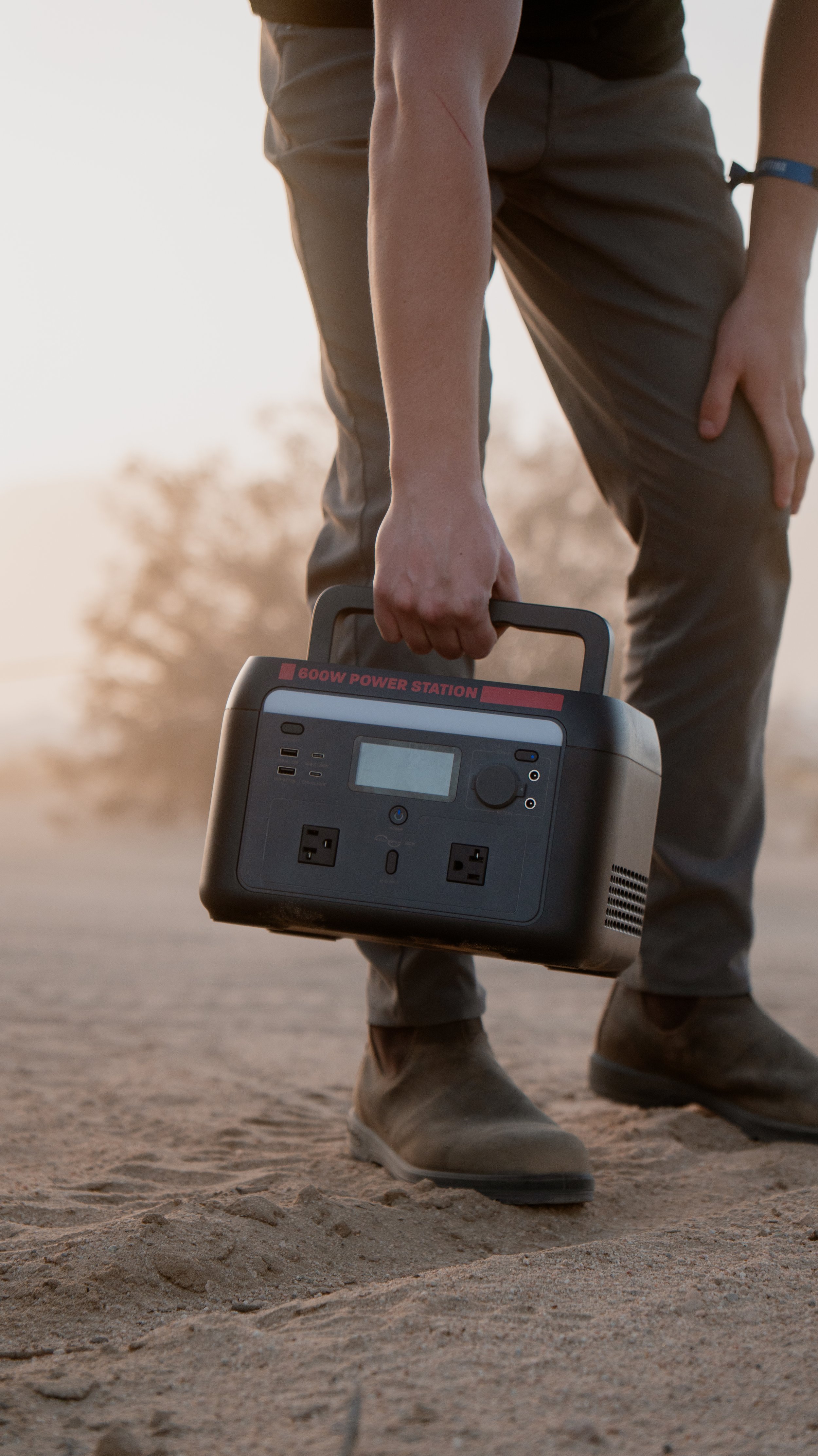 Person holding a portable power station outdoors on sandy ground during sunset.