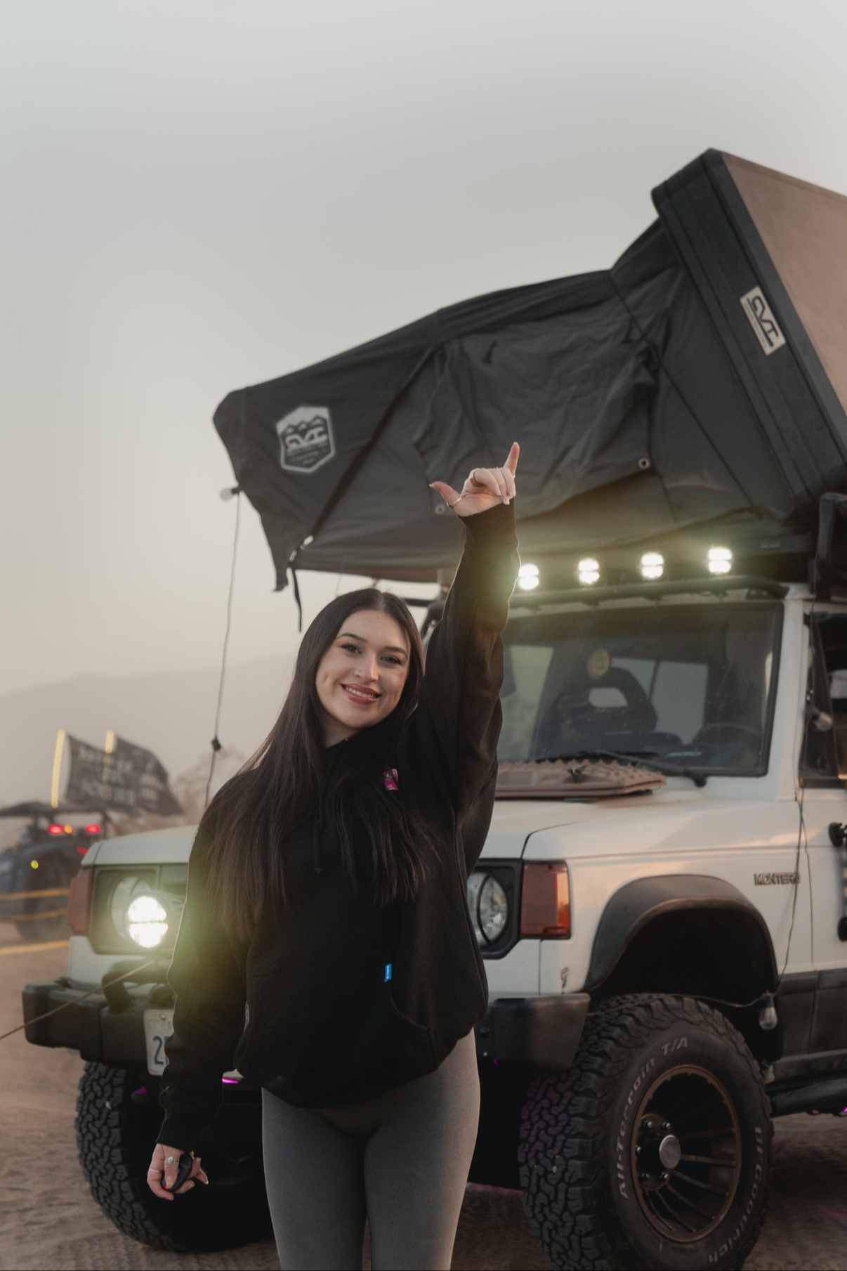 A young woman with long dark hair and a black jacket smiling and raising her right hand with a peace sign in front of an off-road vehicle with a rooftop tent at dusk.