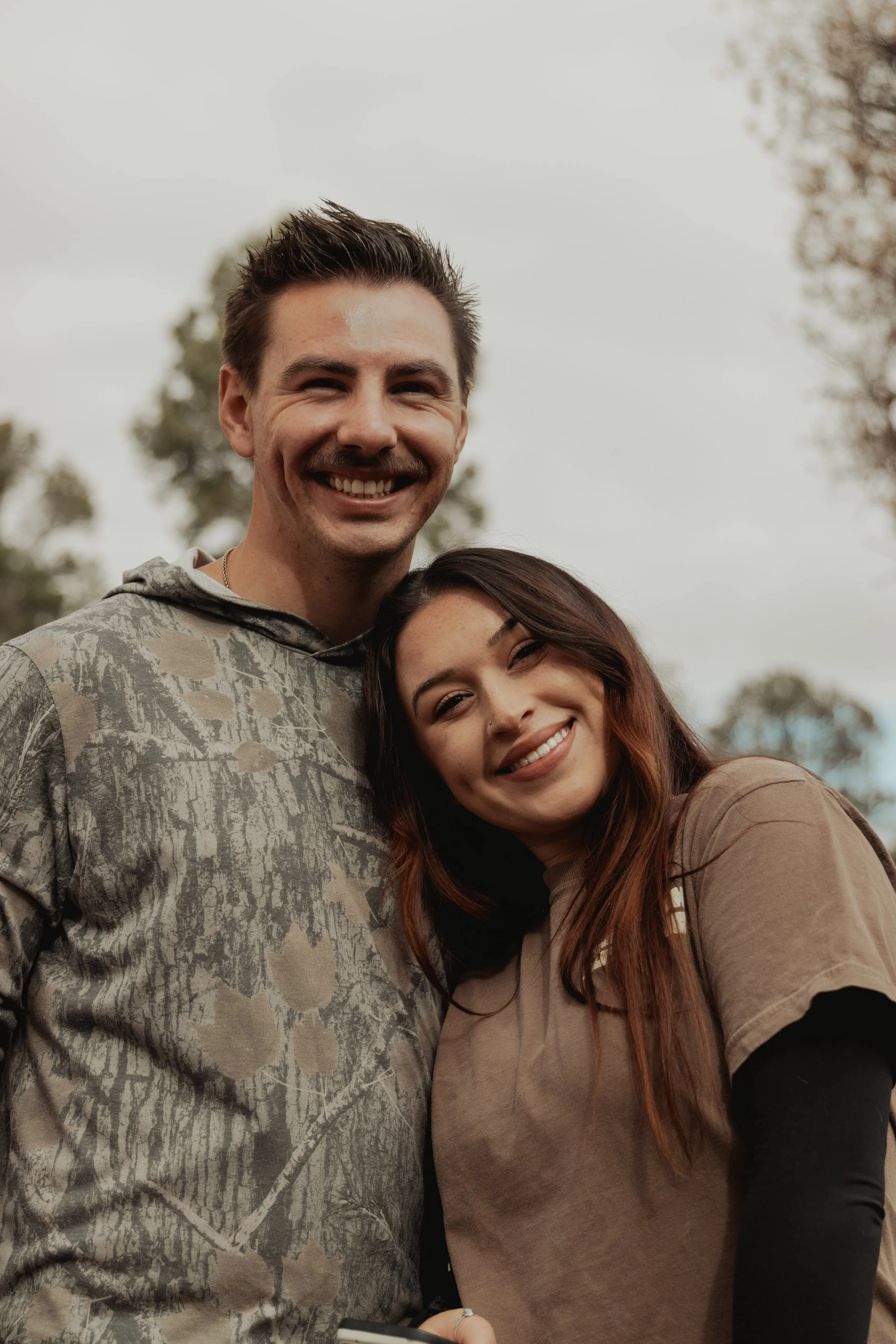 A young couple smiling outdoors, standing close with the man wearing a camouflage hoodie and the woman wearing a beige t-shirt with black long sleeves underneath, against a cloudy sky and trees in the background.