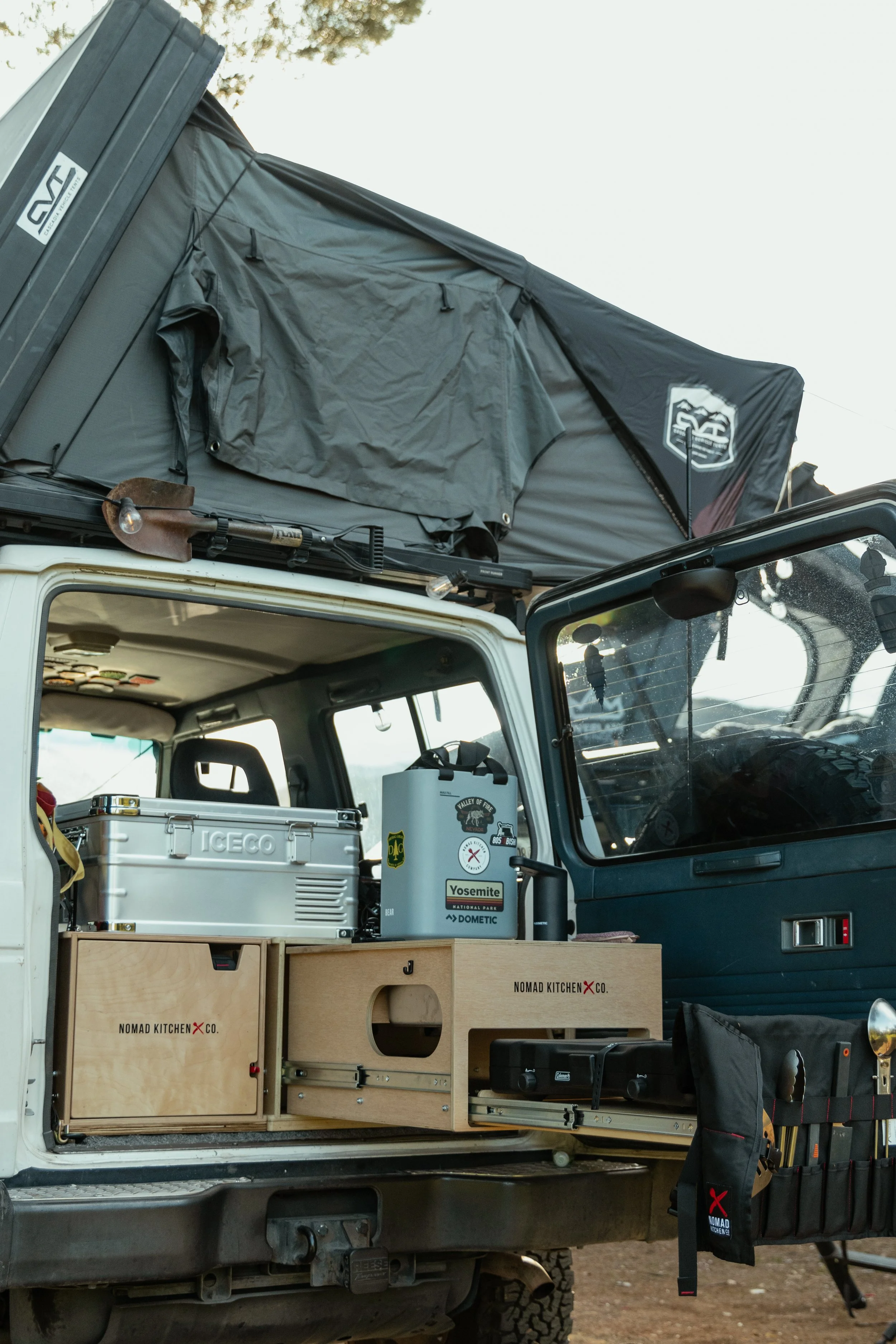 Open vehicle with camping and outdoor gear, including a rooftop tent, kitchen setup with a cooler and water containers, and camping tools, parked outdoors.