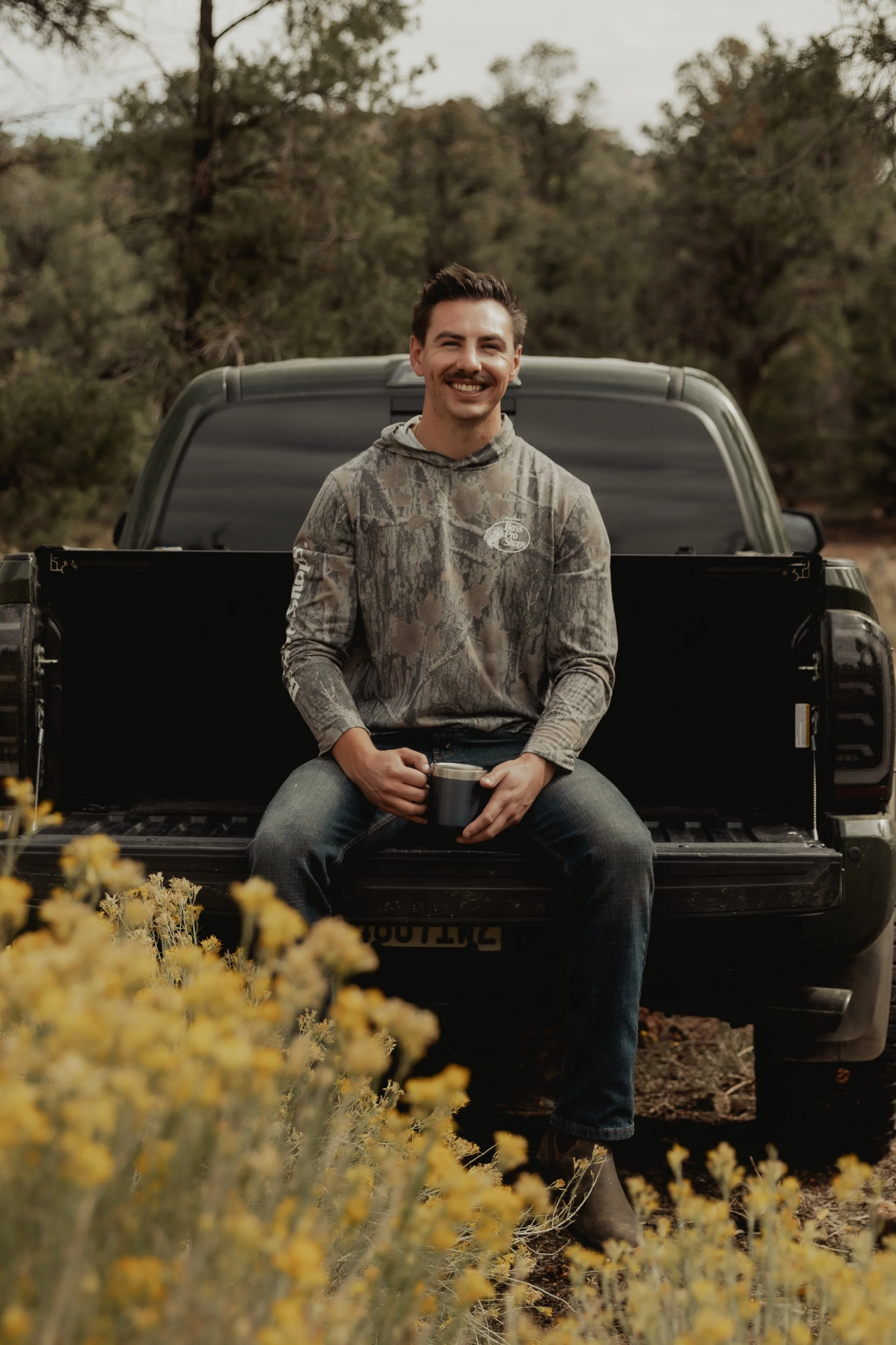 A young man sitting on the tailgate of a black pickup truck in an outdoor setting, holding a metal mug, smiling, with trees and bushes in the background.