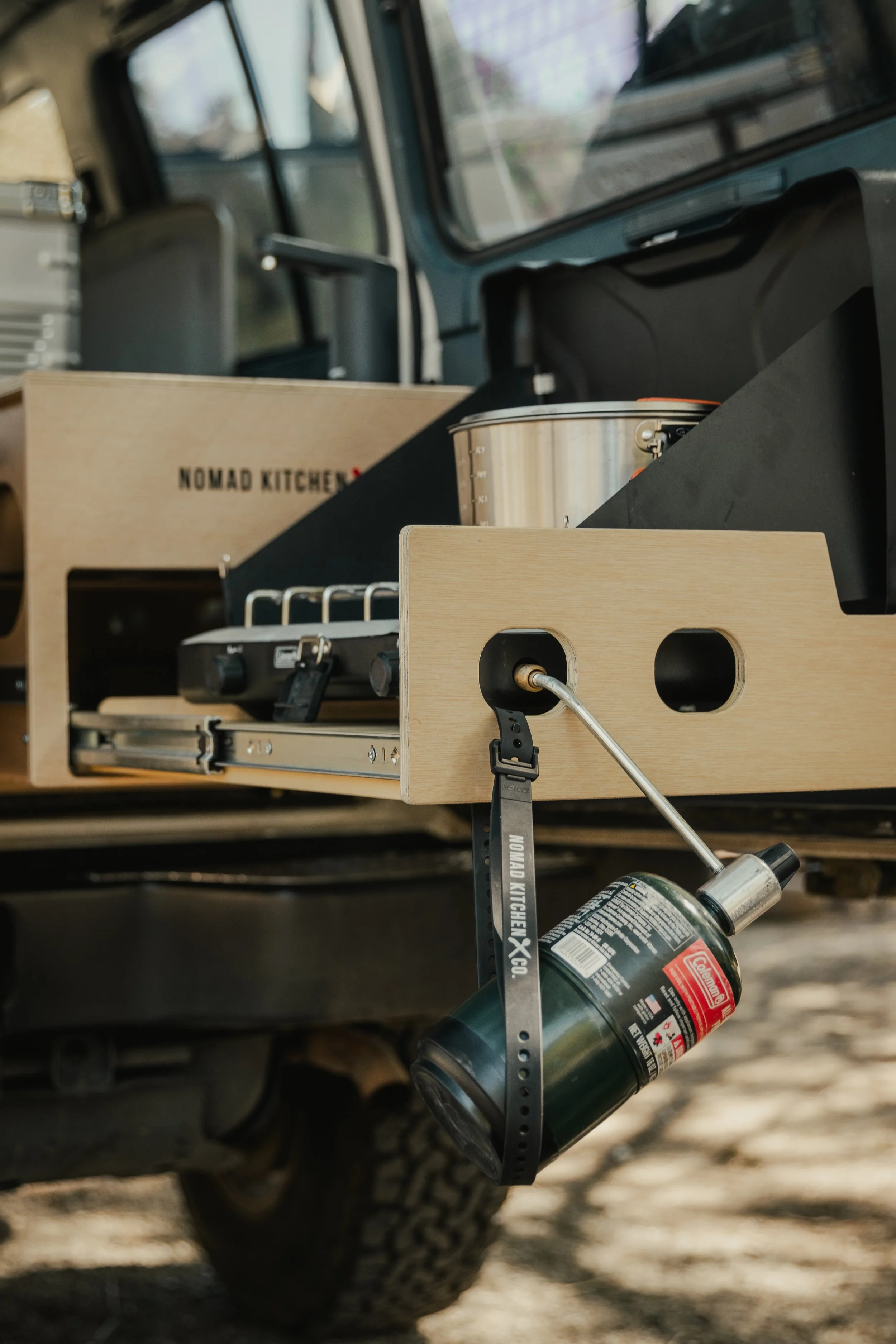 A portable camping stove with a fuel canister mounted under a wooden countertop inside a vehicle, with a cardboard box labeled 'Nomad Kitchen' in the background.