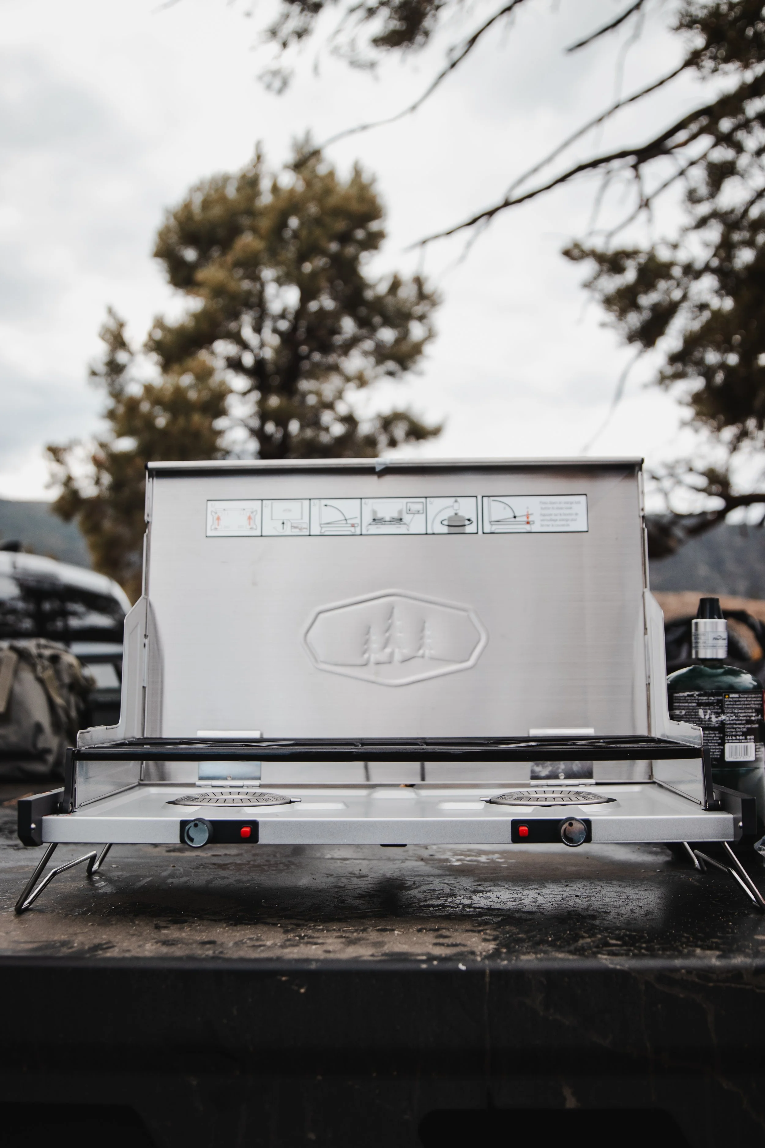 Portable camping stove with two burners on a black table outdoors, with trees and cloudy sky in the background.
